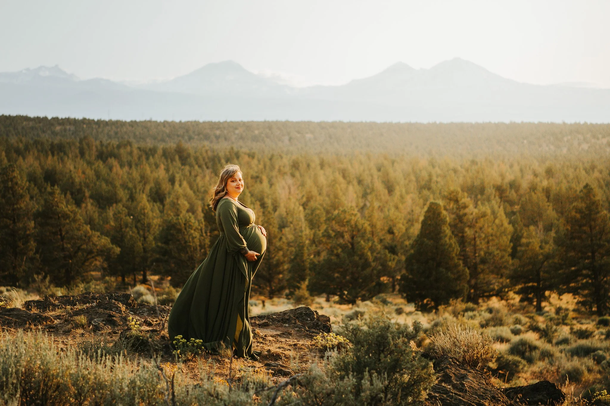 Pregnant woman standing on a rocky outcrop in a desert landscape with a dense forest and mountains in the background at sunset