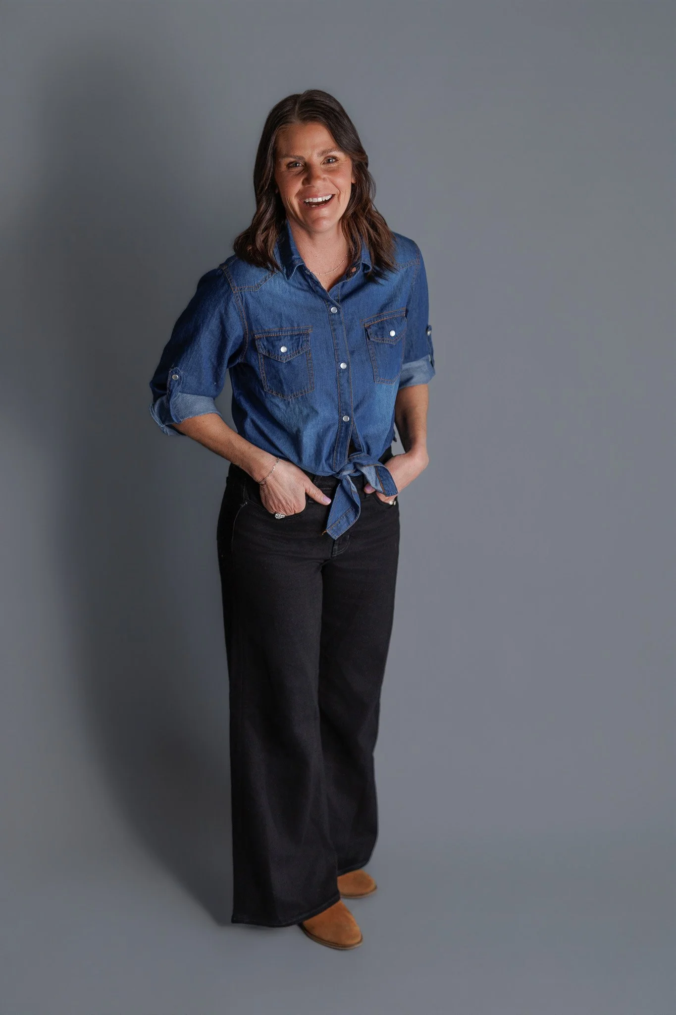 Women in a denim shirt and black pants standing against a gray background