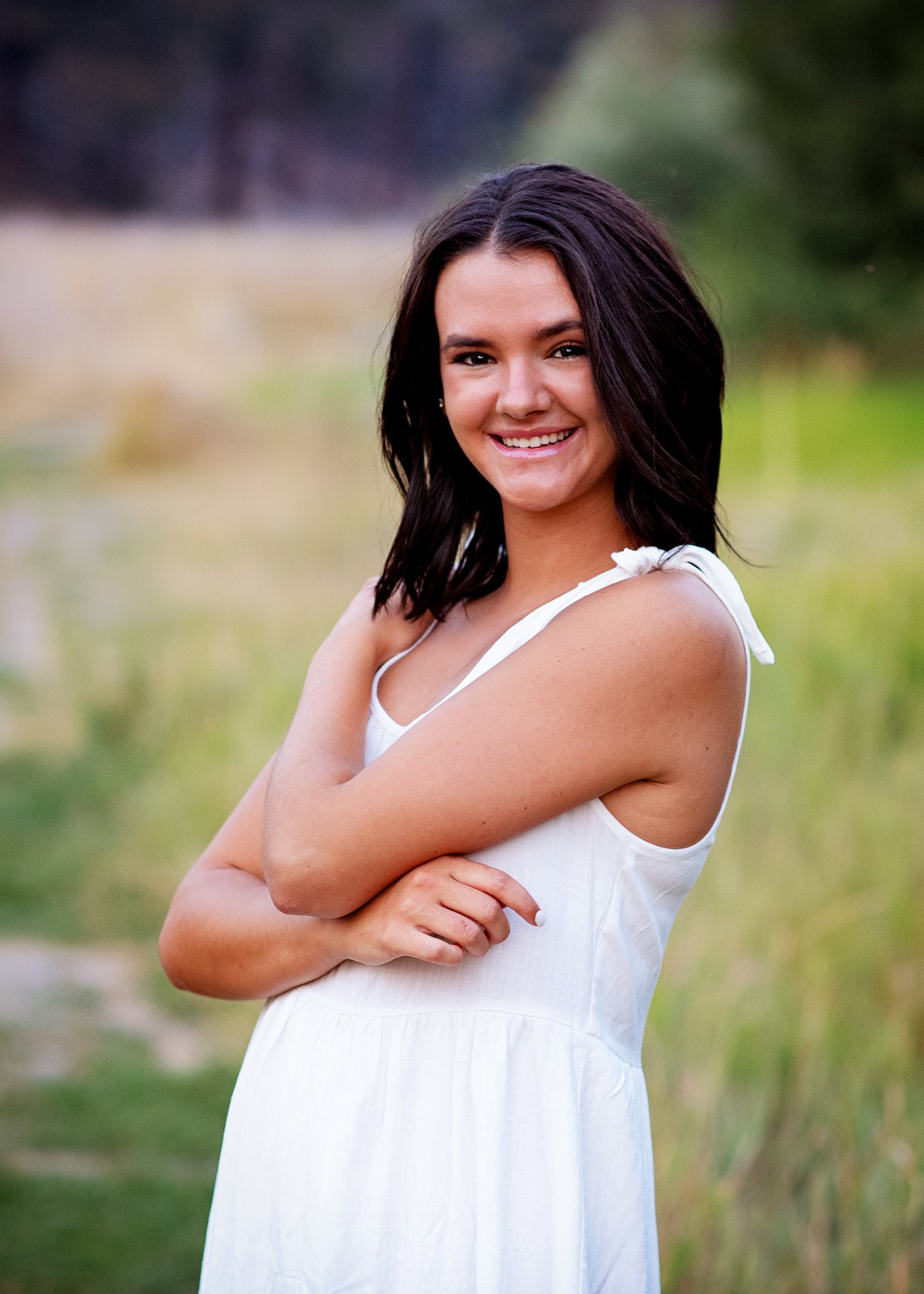 A young woman with dark brown hair and a white sleeveless dress standing outdoors in a grassy area, smiling at the camera.
