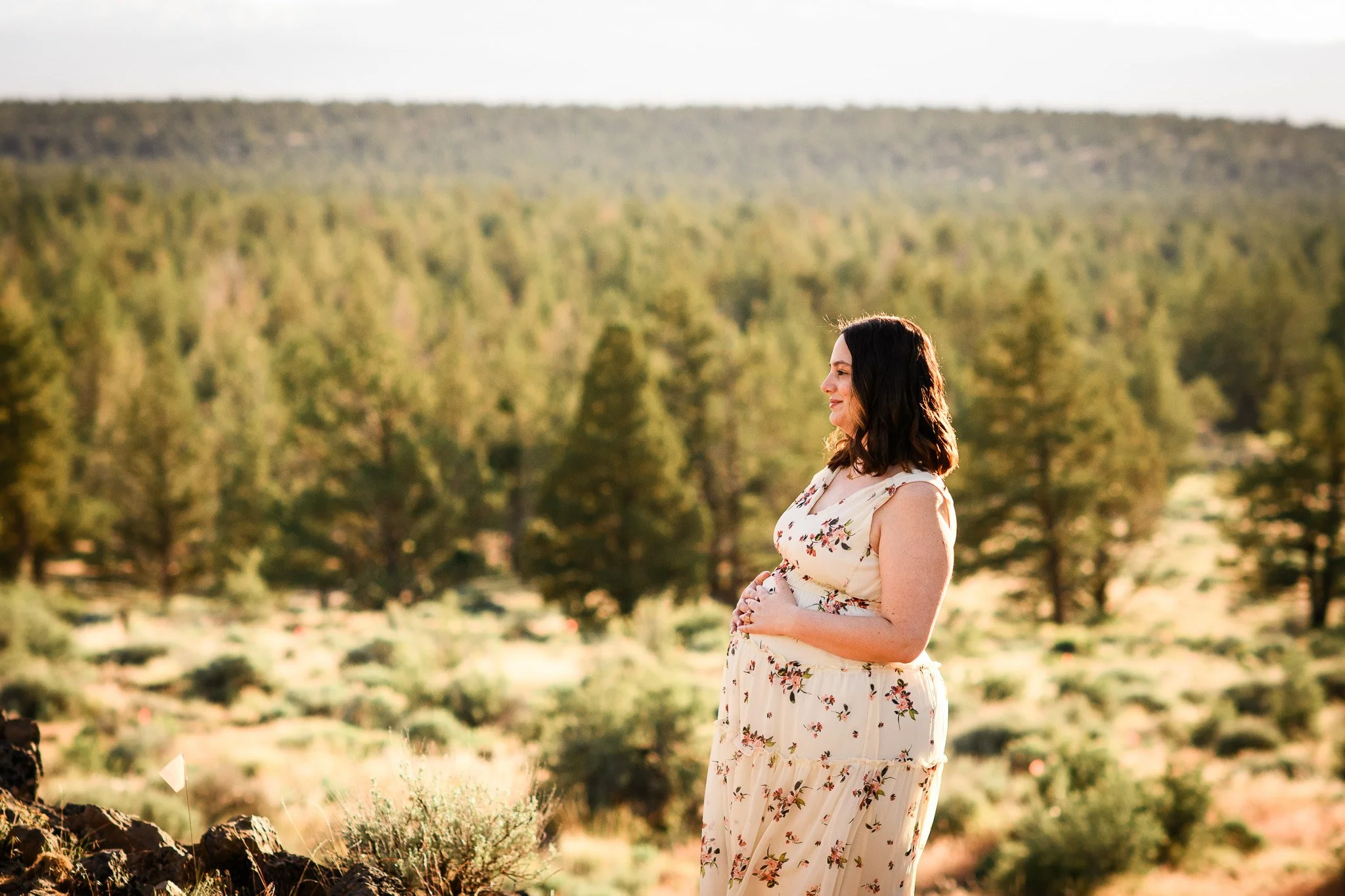 A woman in a floral dress standing outdoors in a desert landscape with trees, facing to the right, during daytime.