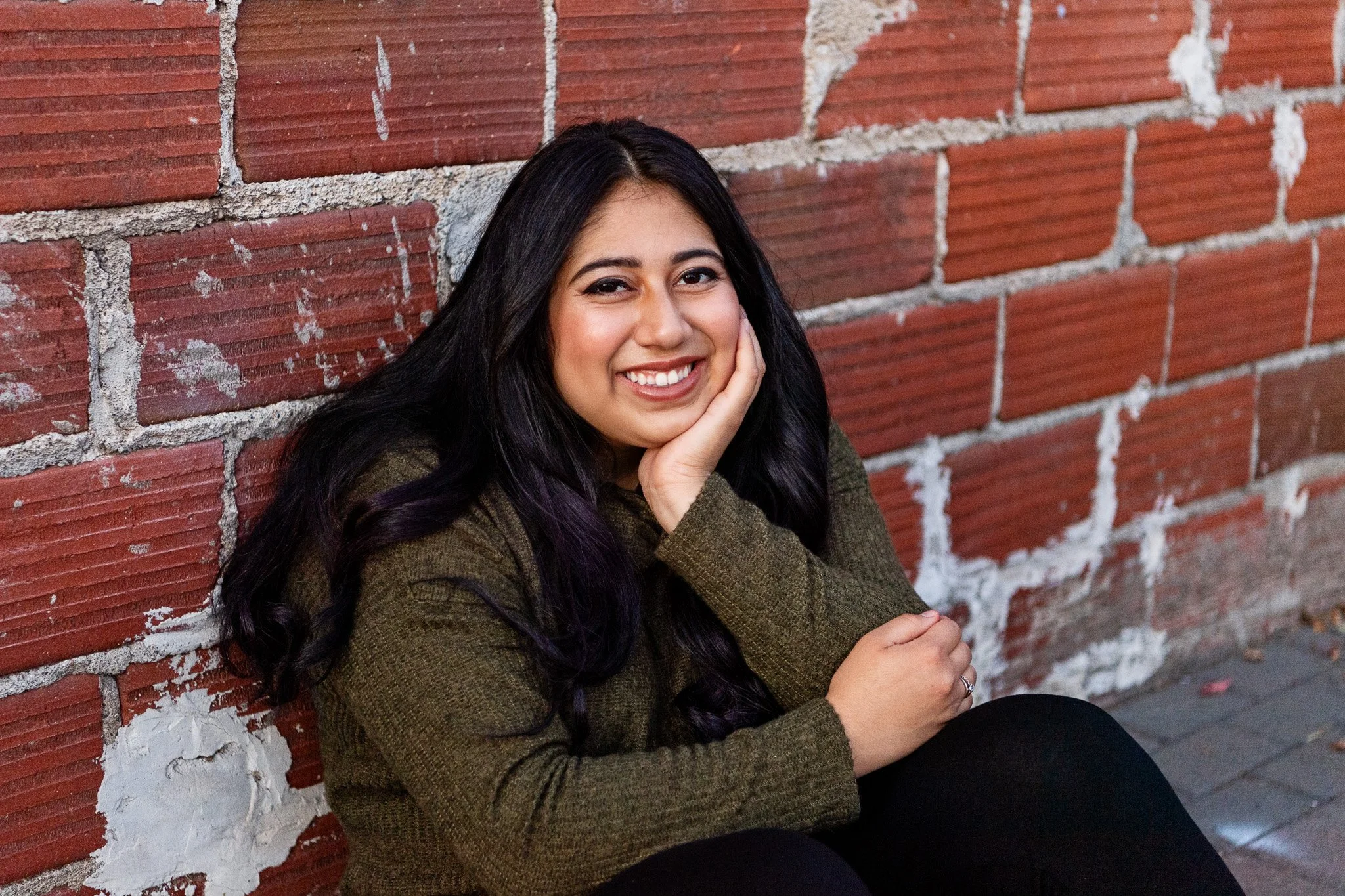 A young woman with long dark hair, smiling, sitting against a brick wall, resting her head on her hand, wearing a brown sweater.
