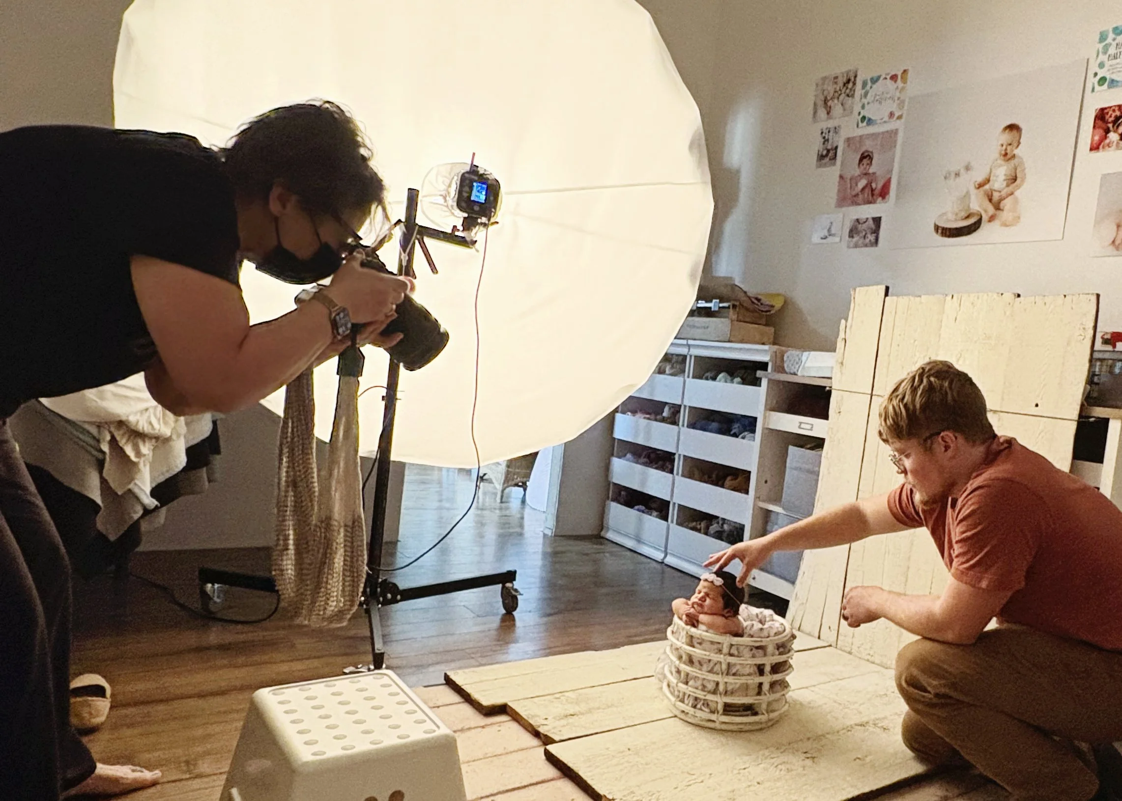Photographer takes pictures of a newborn baby sitting in a basket on a wooden platform during a photo shoot with a large umbrella reflector and a man adjusting the baby's head.