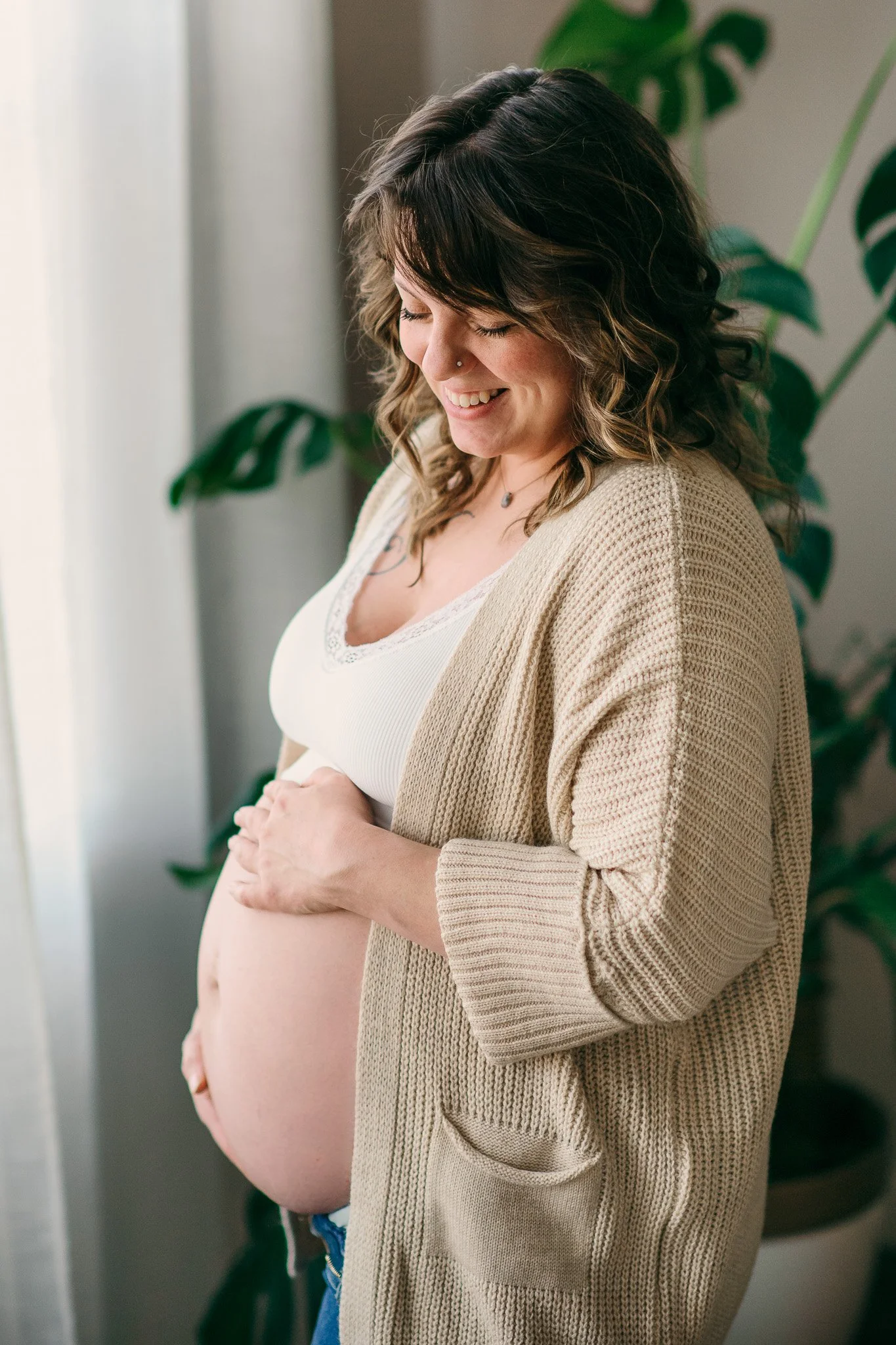 A pregnant woman with curly brown hair smiling and looking down, wearing a white top and beige cardigan, standing near a window with green plants in the background.
