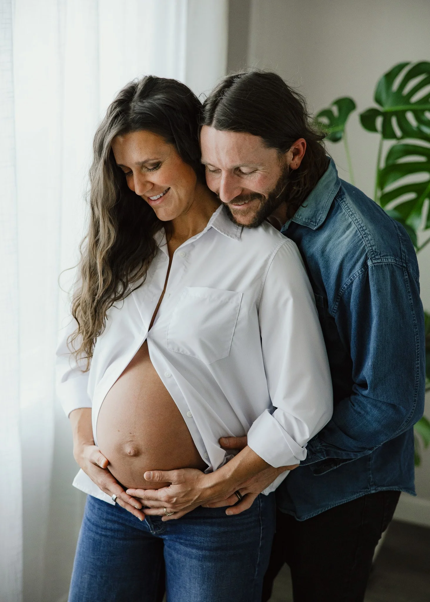 A pregnant woman with long dark hair in a white shirt, smiling, holds her belly with her hands, while a man with long dark hair and beard, in a denim shirt, embraces her from behind and smiles, in a well-lit room with a large green plant in the backg