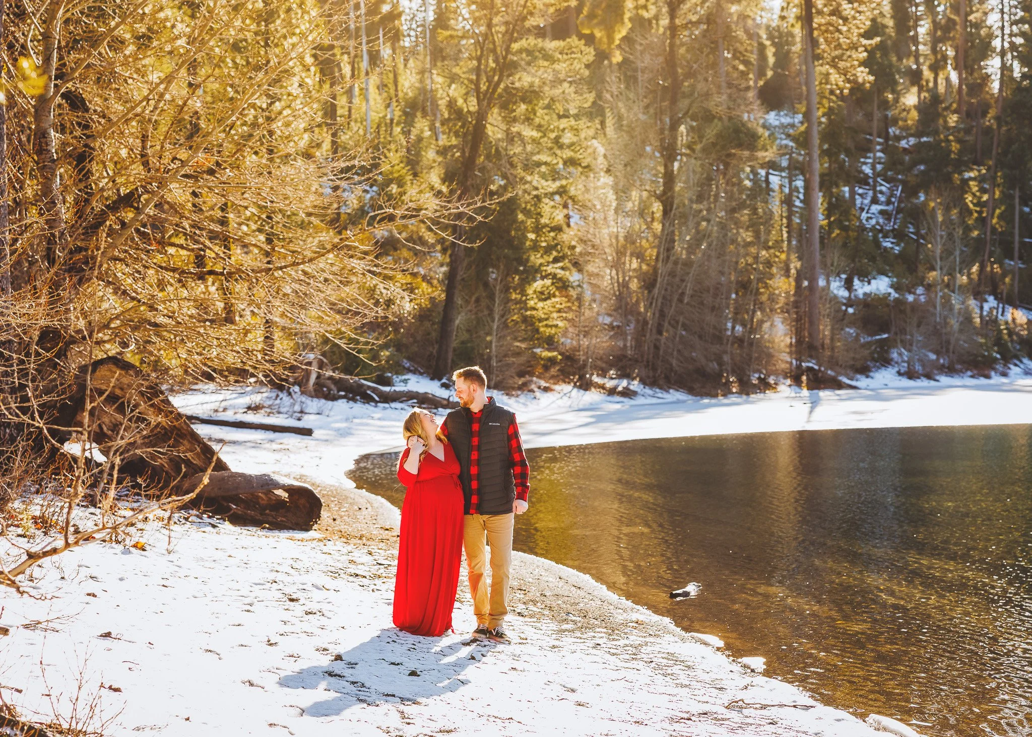 A young couple dressed in red and black, standing on a snowy riverbank, embracing and looking at each other in a sunlit winter forest with trees and snow in the background.