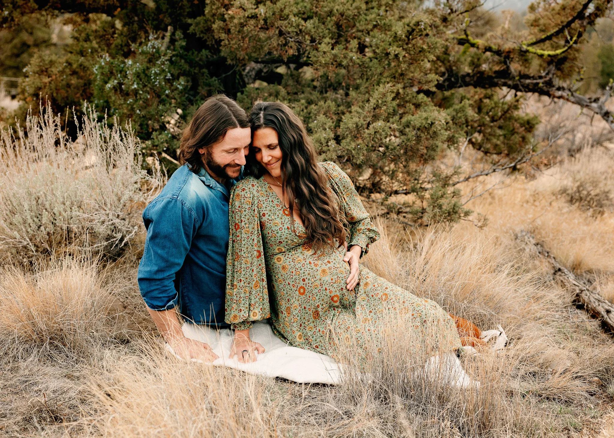 A couple sitting closely outdoors in a grassy area with shrubs and trees, with the woman appearing pregnant, both smiling and looking down.