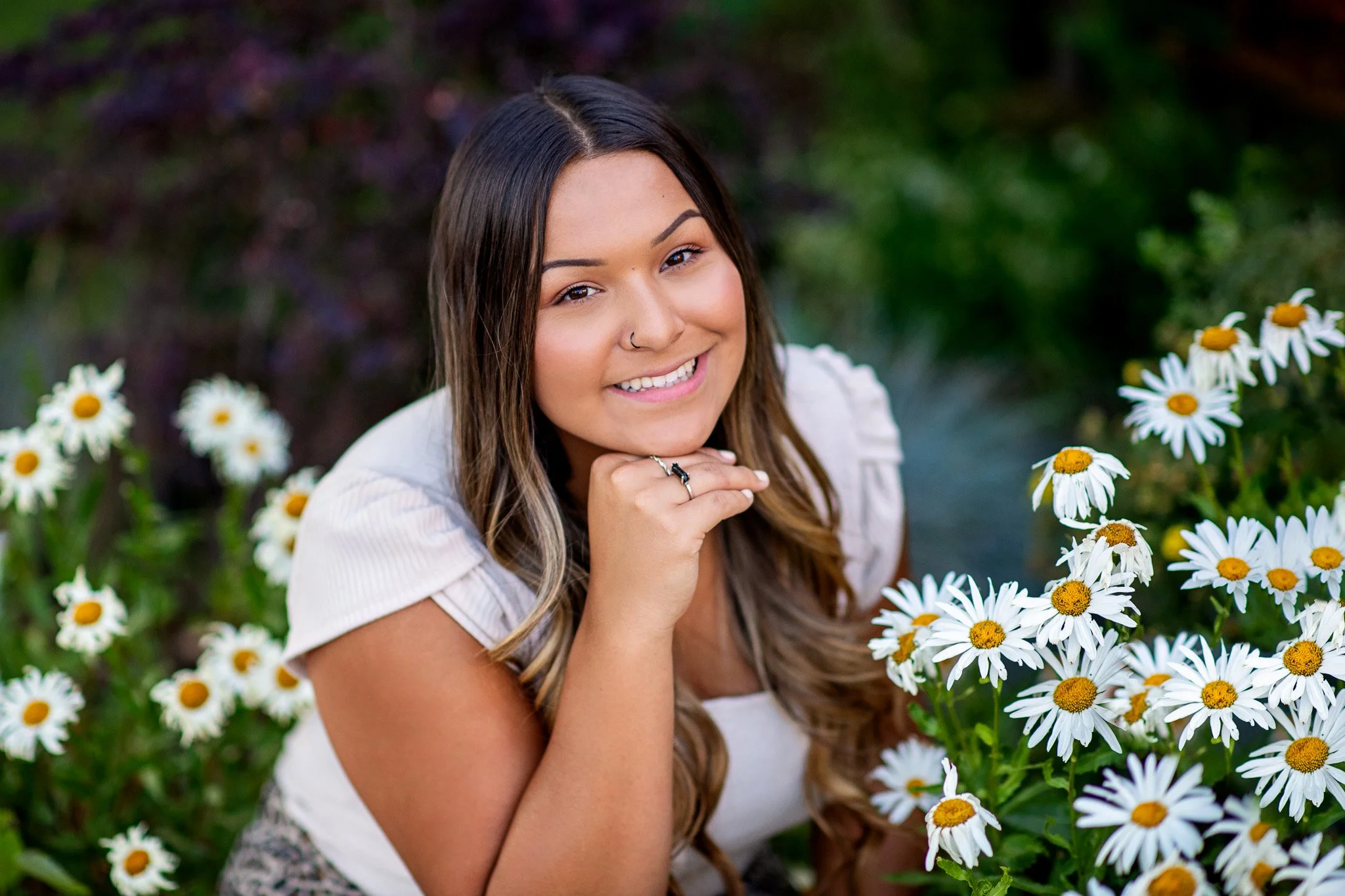 A young woman with long, wavy brown hair and light skin, smiling, resting her chin on her hand, surrounded by white daisies outdoors.