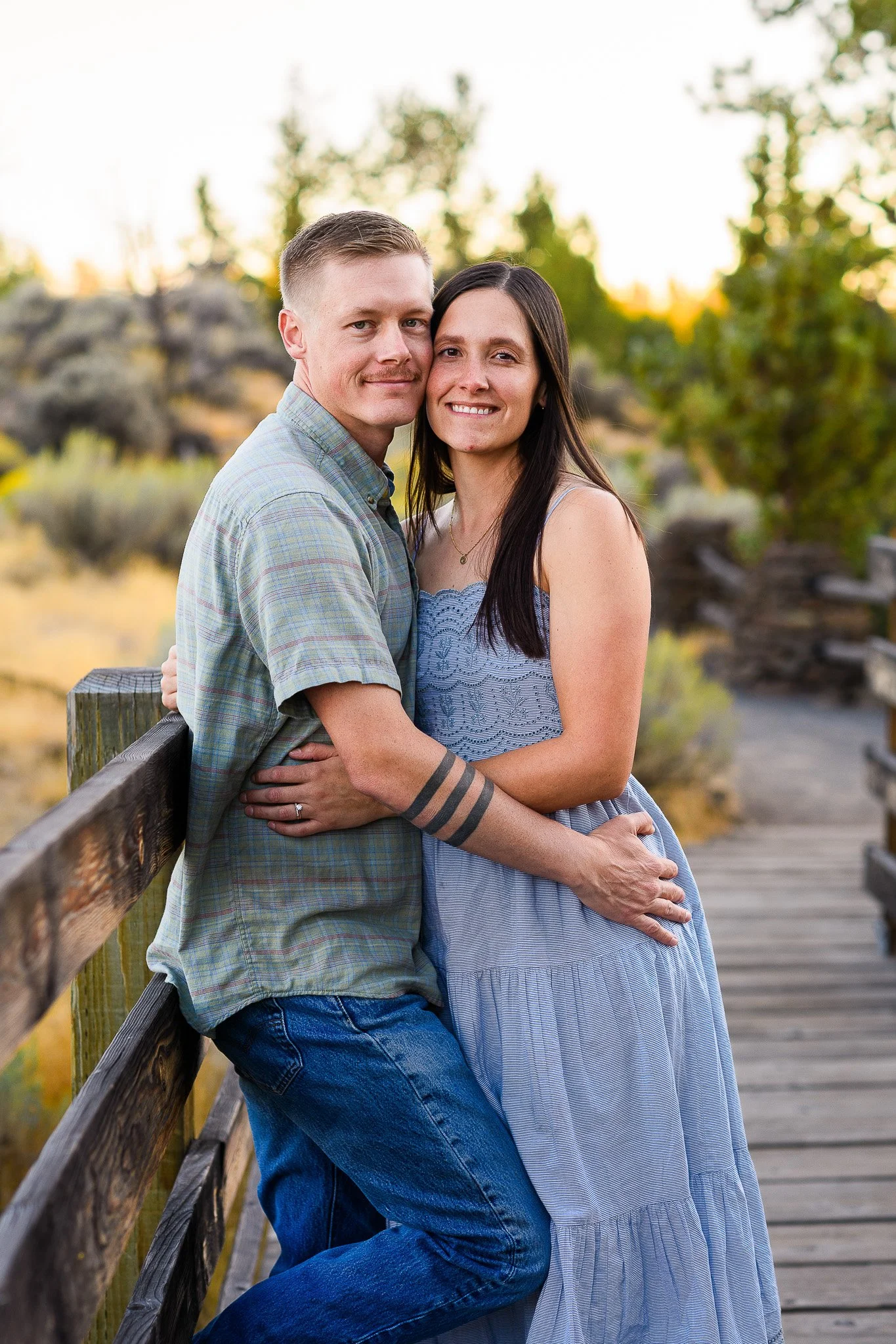 A couple embraces on a wooden bridge outdoors, smiling at the camera with a background of trees and a sunset sky.