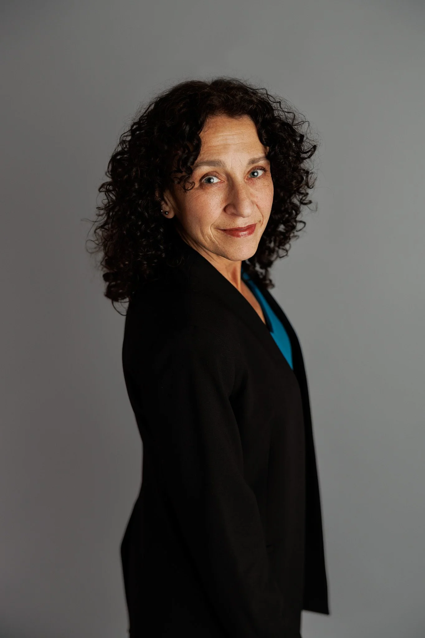 Portrait of a middle-aged woman with curly dark hair, wearing a black blazer and a blue top, standing against a gray background and smiling slightly.