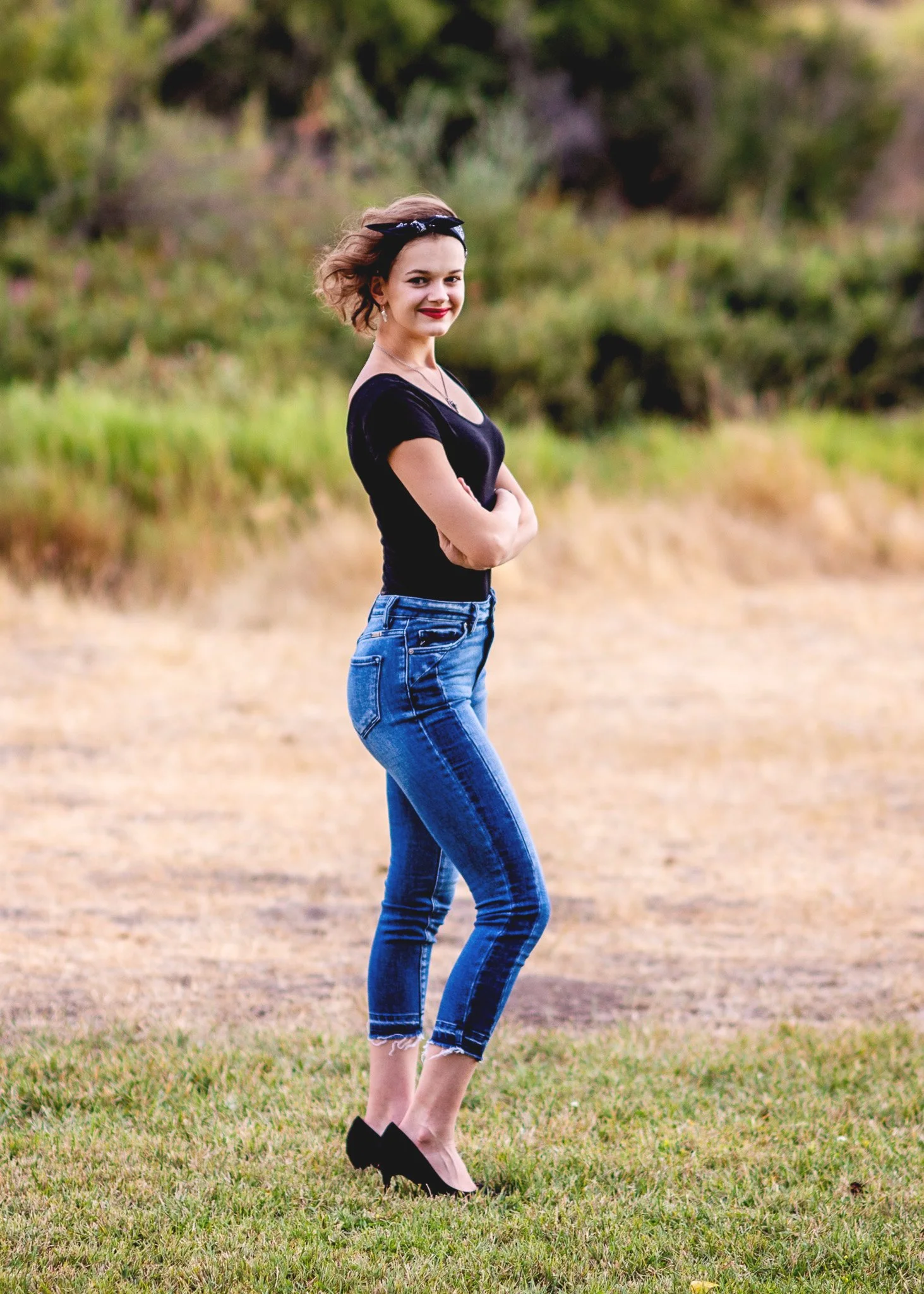 A woman standing outdoors on grass, smiling, wearing a black t-shirt, blue jeans, black shoes, and a headband, with trees and a dirt path in the background.