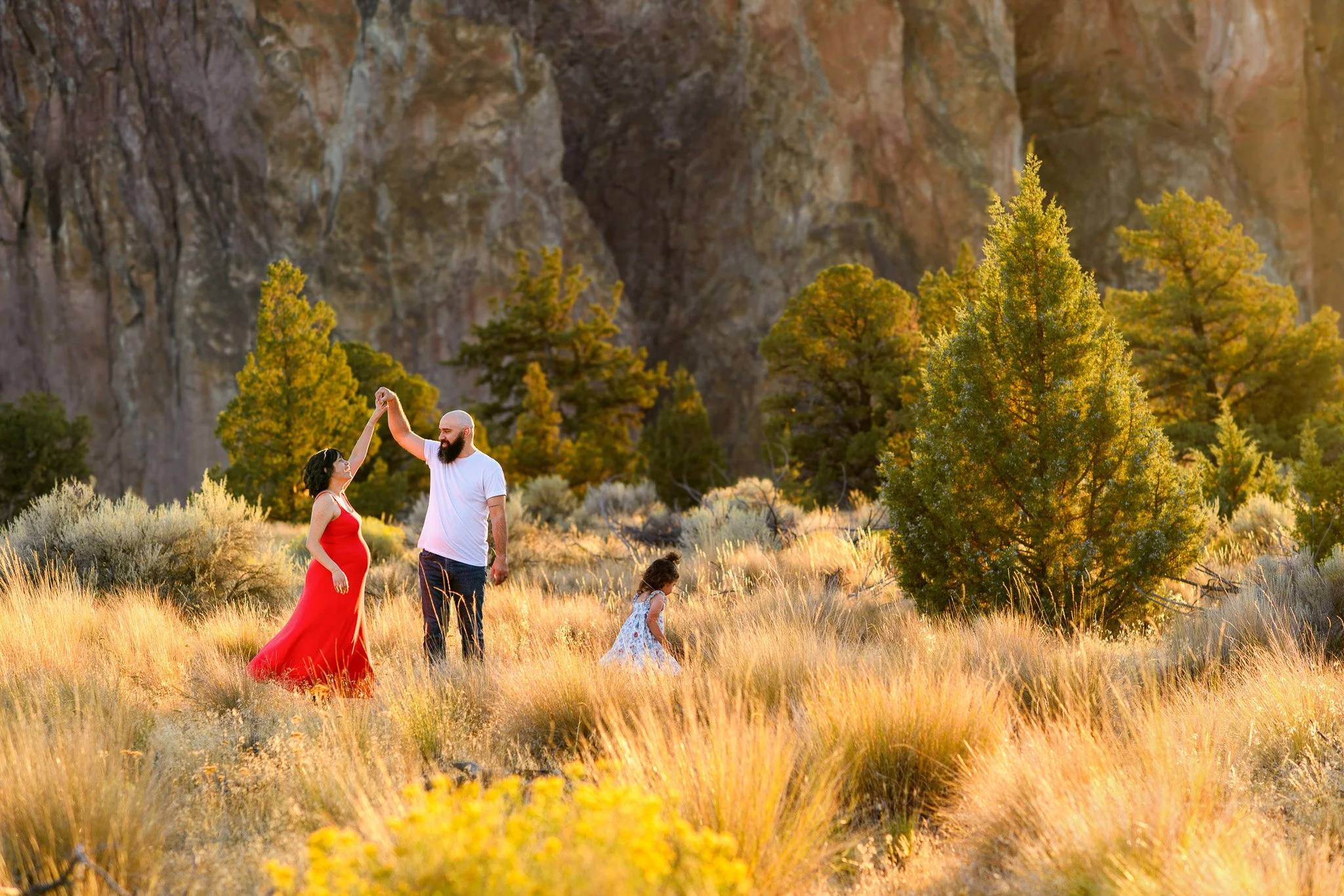 A couple dancing in a grassy field with a young girl nearby, surrounded by trees and rocky cliffs during sunset.