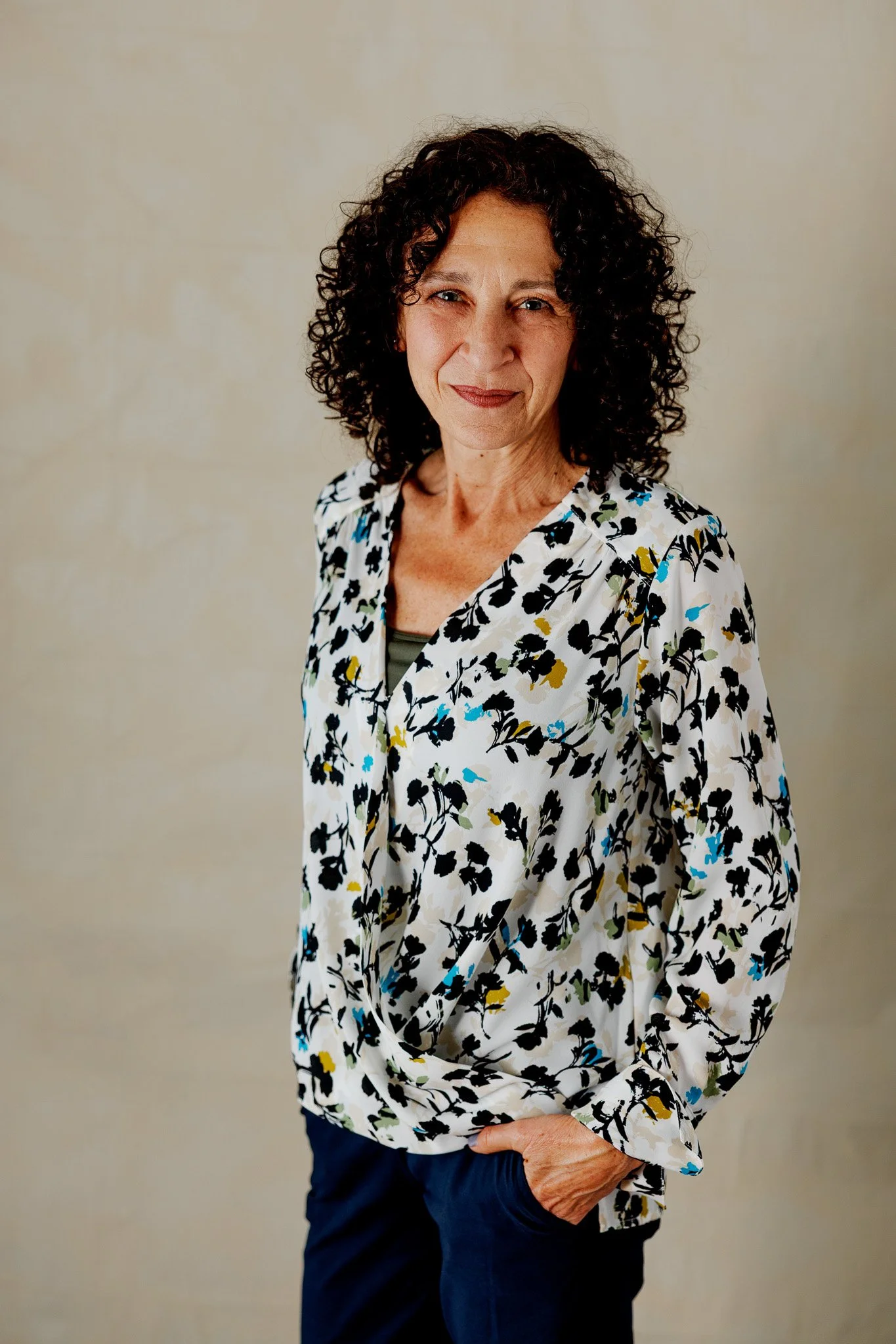 A middle-aged woman with curly dark hair, wearing a patterned blouse with blue, yellow, and black accents, and dark pants, standing against a plain, light-colored background, looking at the camera with a slight smile.