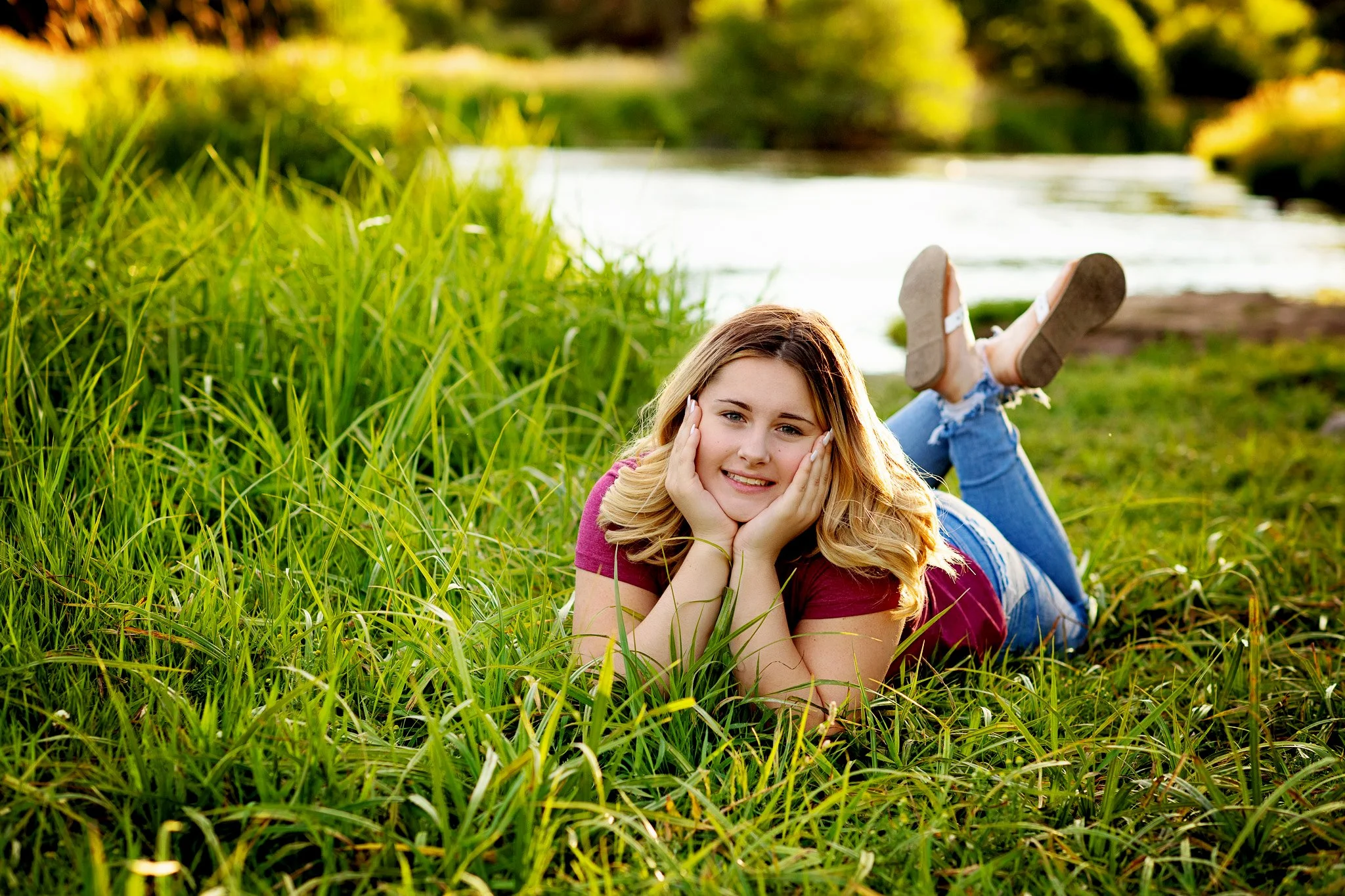 A young woman lying on her stomach in green grass near a river, smiling with her hands on her cheeks, wearing a pink shirt, ripped jeans, and sandals, with trees and water in the background.