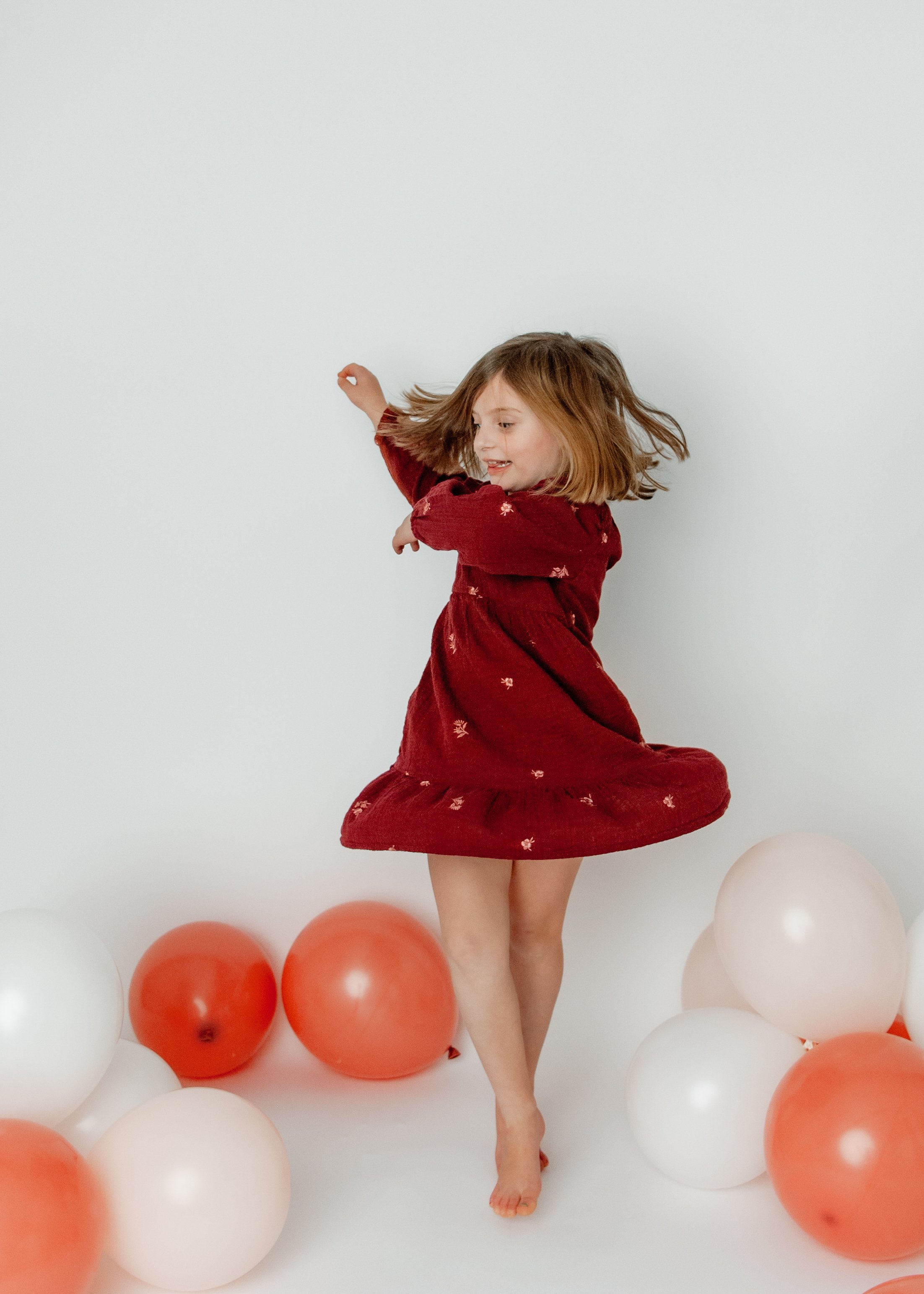 A young girl in a red dress is twirling with balloons around her, against a white background.
