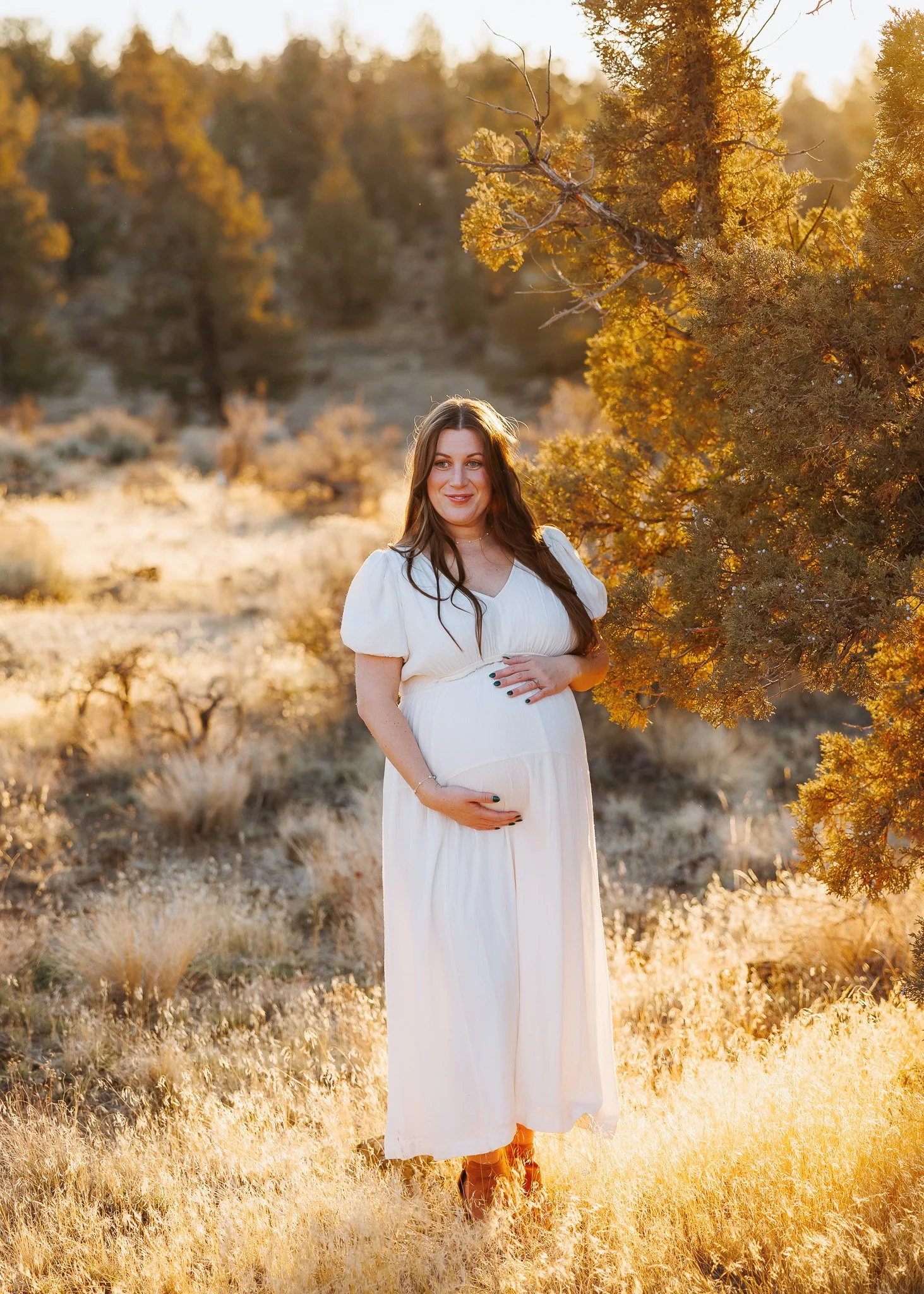 A pregnant woman in a white dress standing outdoors in a desert landscape with yellowish grass and trees during sunset, gently holding her belly and smiling.