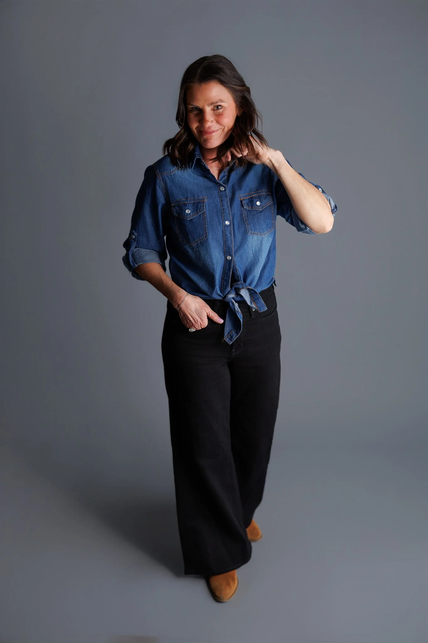Woman in denim shirt and black pants posing on gray background