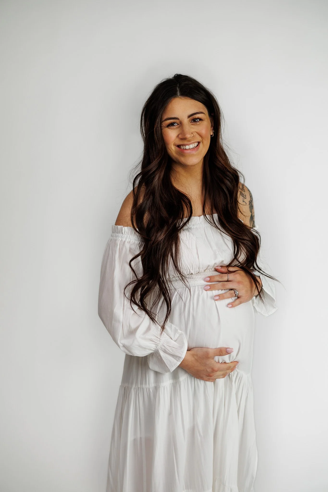 A pregnant woman with long brown hair smiling and holding her belly, wearing a white off-shoulder dress, standing against a plain white background.