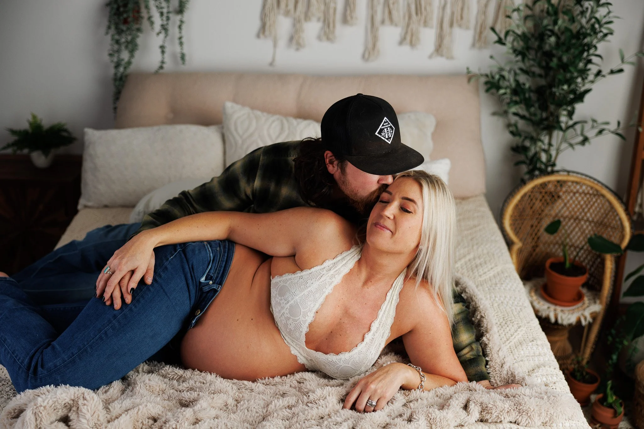 A man wearing a black cap and plaid shirt is kissing a woman with blonde hair who is lying on a bed wearing a white lace top and blue jeans. The room has a beige headboard, white pillows, plants, and bohemian-style decor.