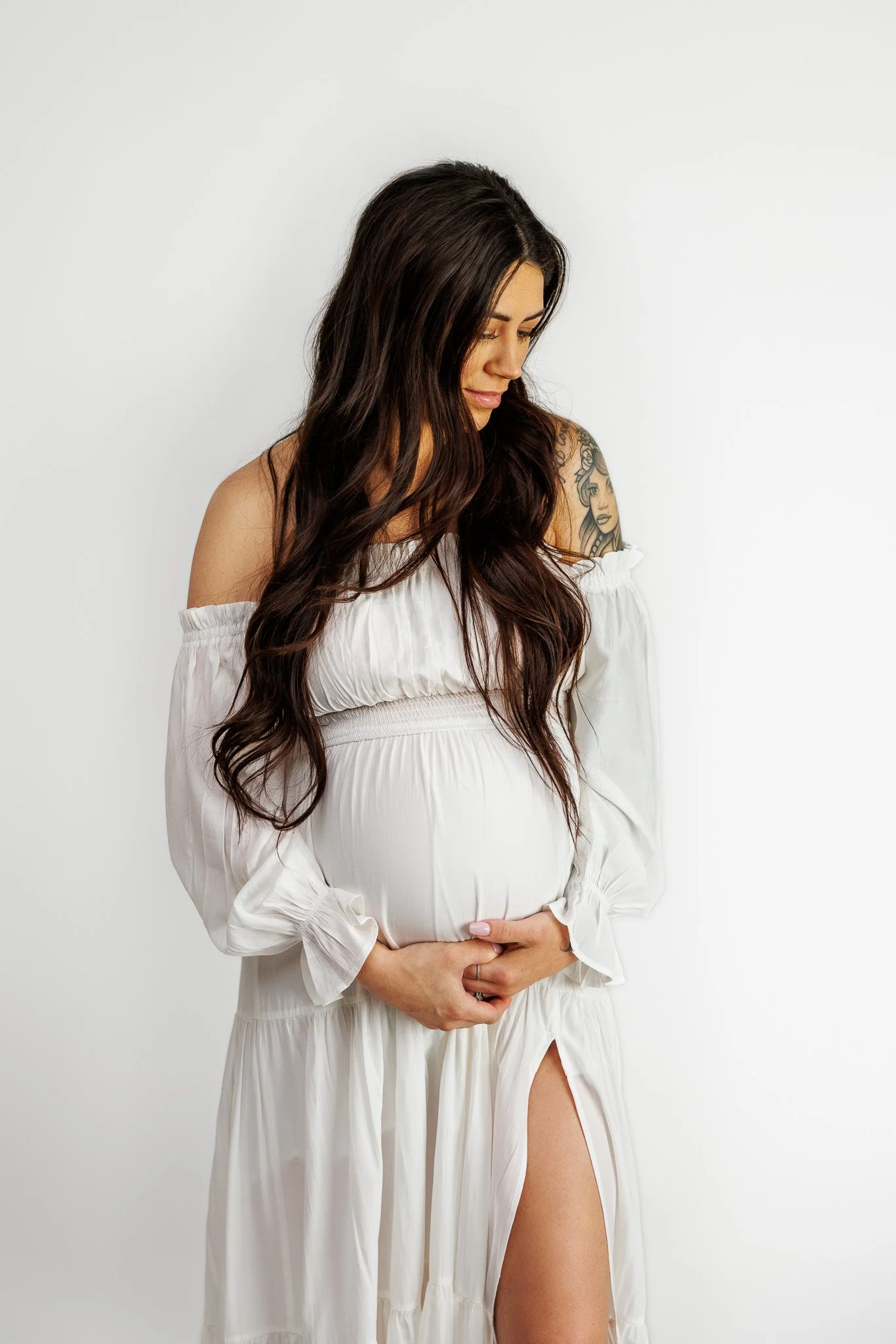 Pregnant woman with long brown hair, wearing a white dress with off-shoulder sleeves, standing against a plain white background, holding her belly, with a tattoo on her shoulder.