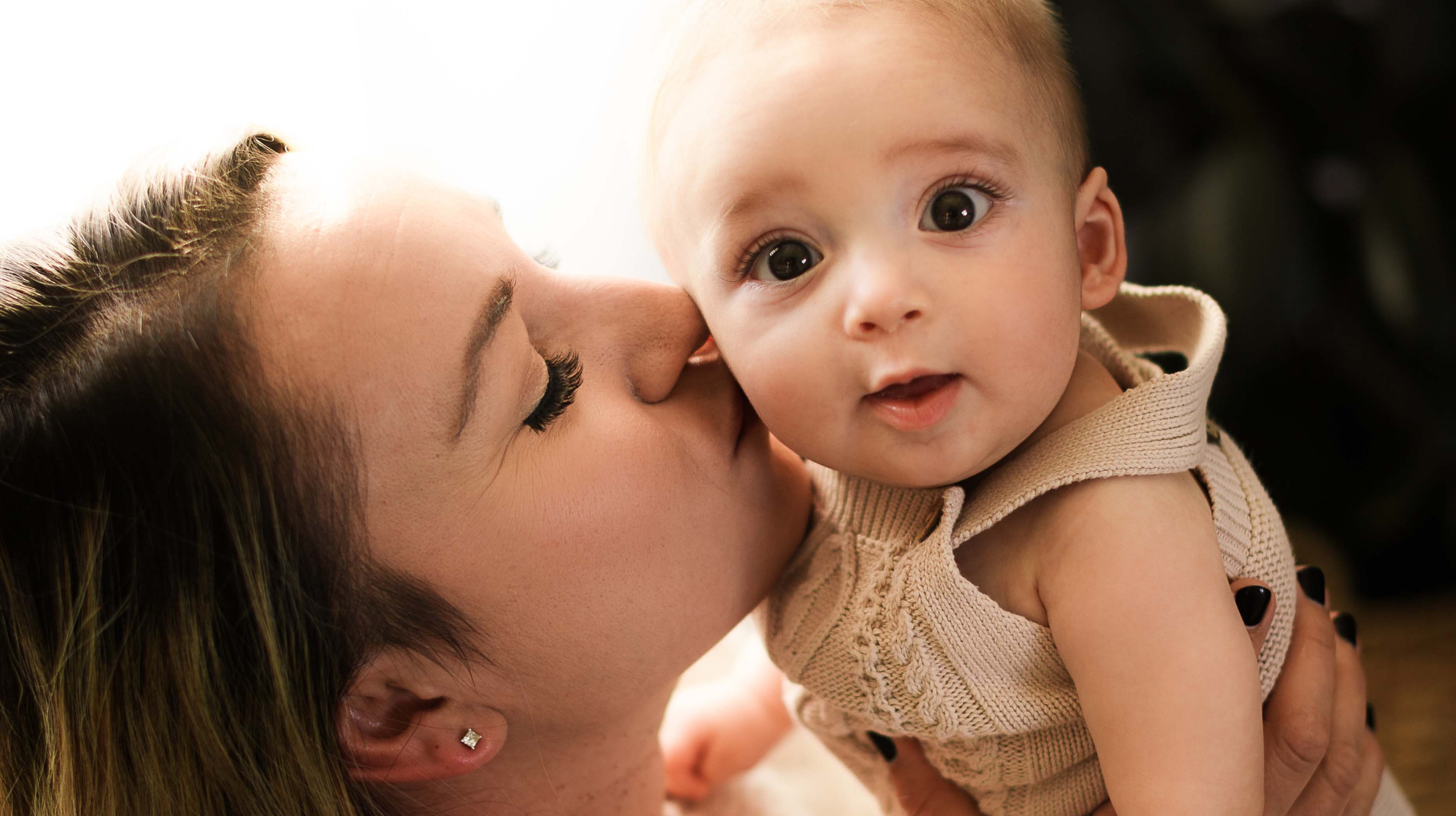 A woman is kissing a baby on the cheek. The baby is looking at the camera with wide eyes and an open mouth. The woman has closed eyes and holds the baby close.