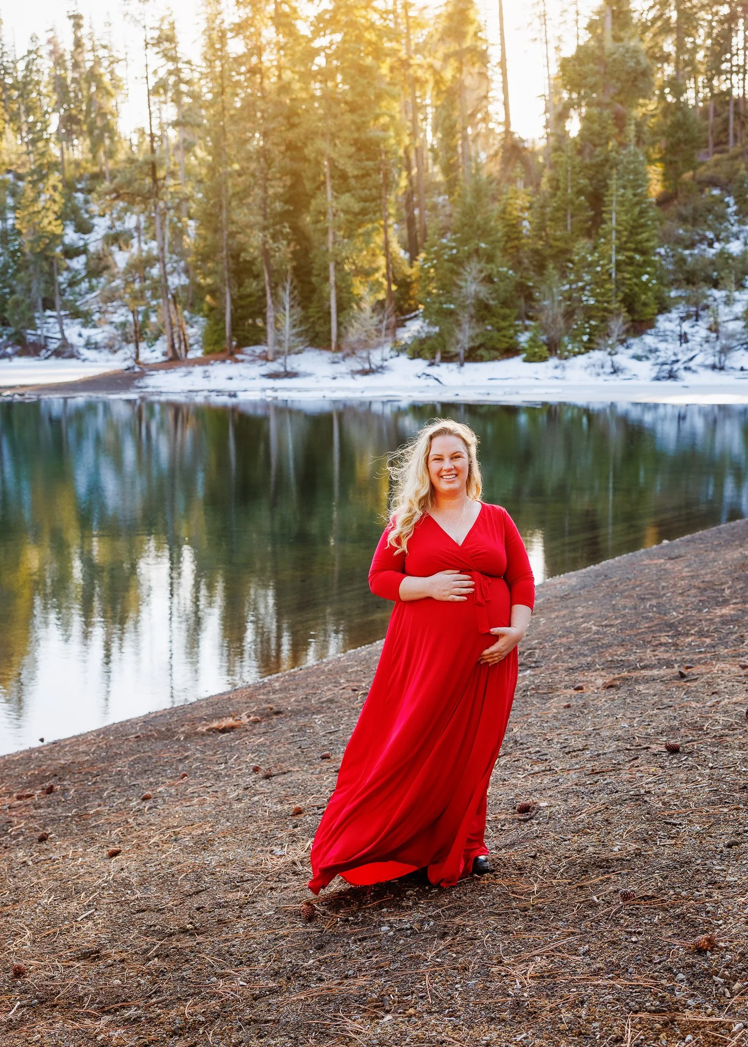 A pregnant woman in a long red dress standing on a dirt path beside a lake, with a snowy forest and pine trees in the background, during sunset.