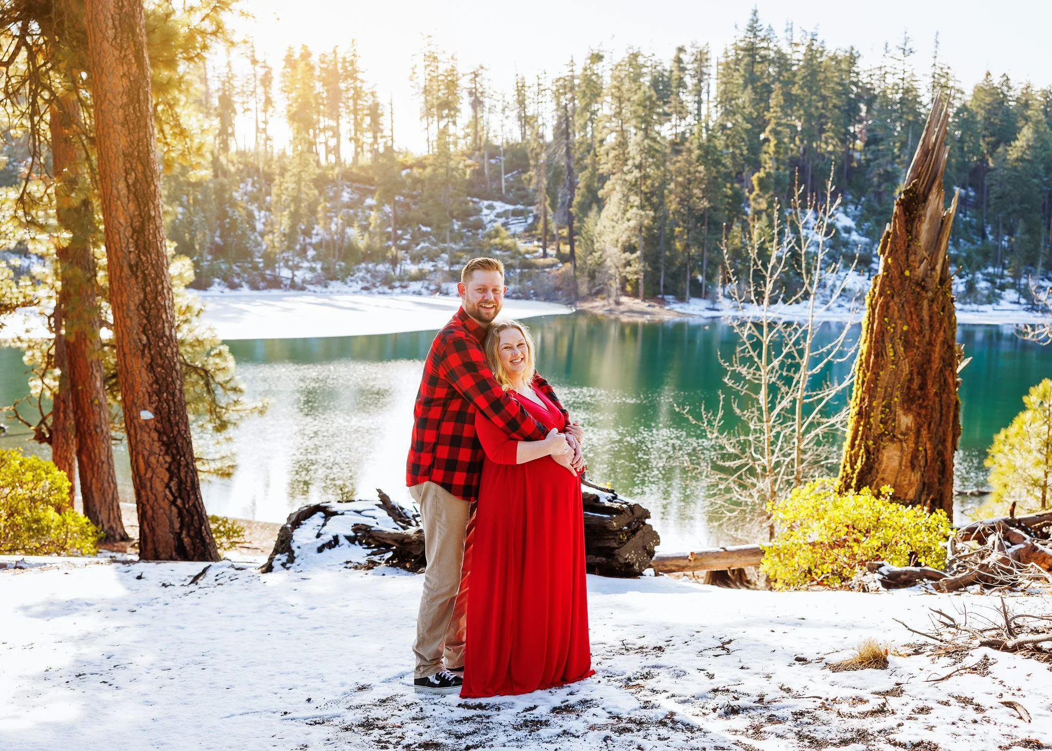 A happy couple stands on snow-covered ground near a lake, with pine trees and a forested hillside in the background. The man is wearing a red and black plaid shirt and beige pants, and the woman is dressed in a long red dress. They are smiling and em