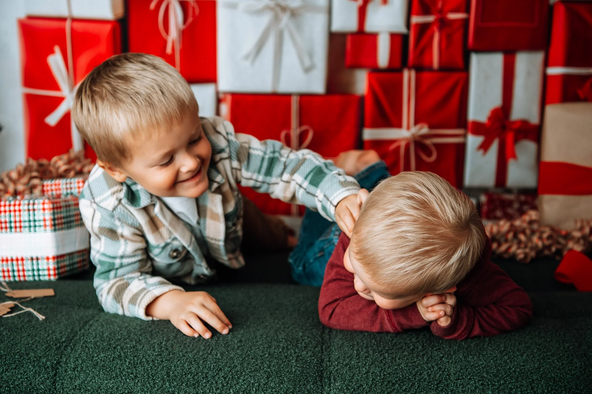 Two young boys playing and laughing in front of a backdrop of Christmas gift boxes, with one boy pinning the other down on a green surface.