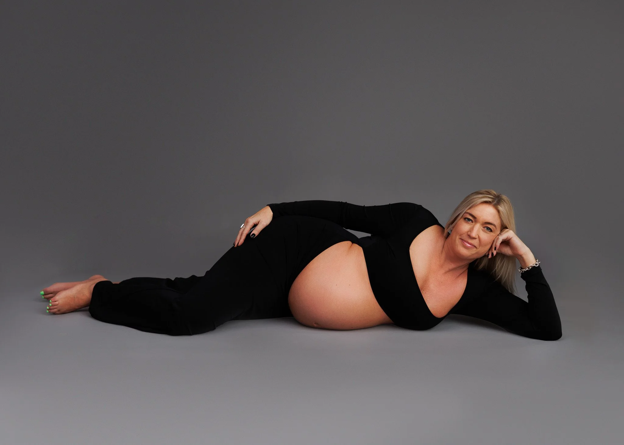 Pregnant woman in black clothing lying on her side on a gray background, smiling and posing for the camera.