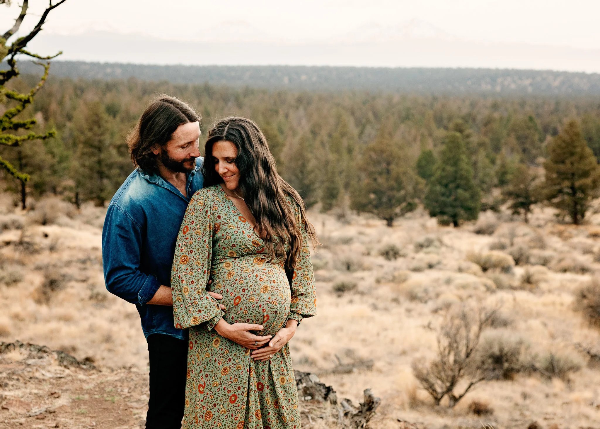 A pregnant woman in a floral green dress stands outdoors with a man in a blue shirt behind her. Both are smiling and looking at each other, with the man's hand on her belly in a desert landscape.