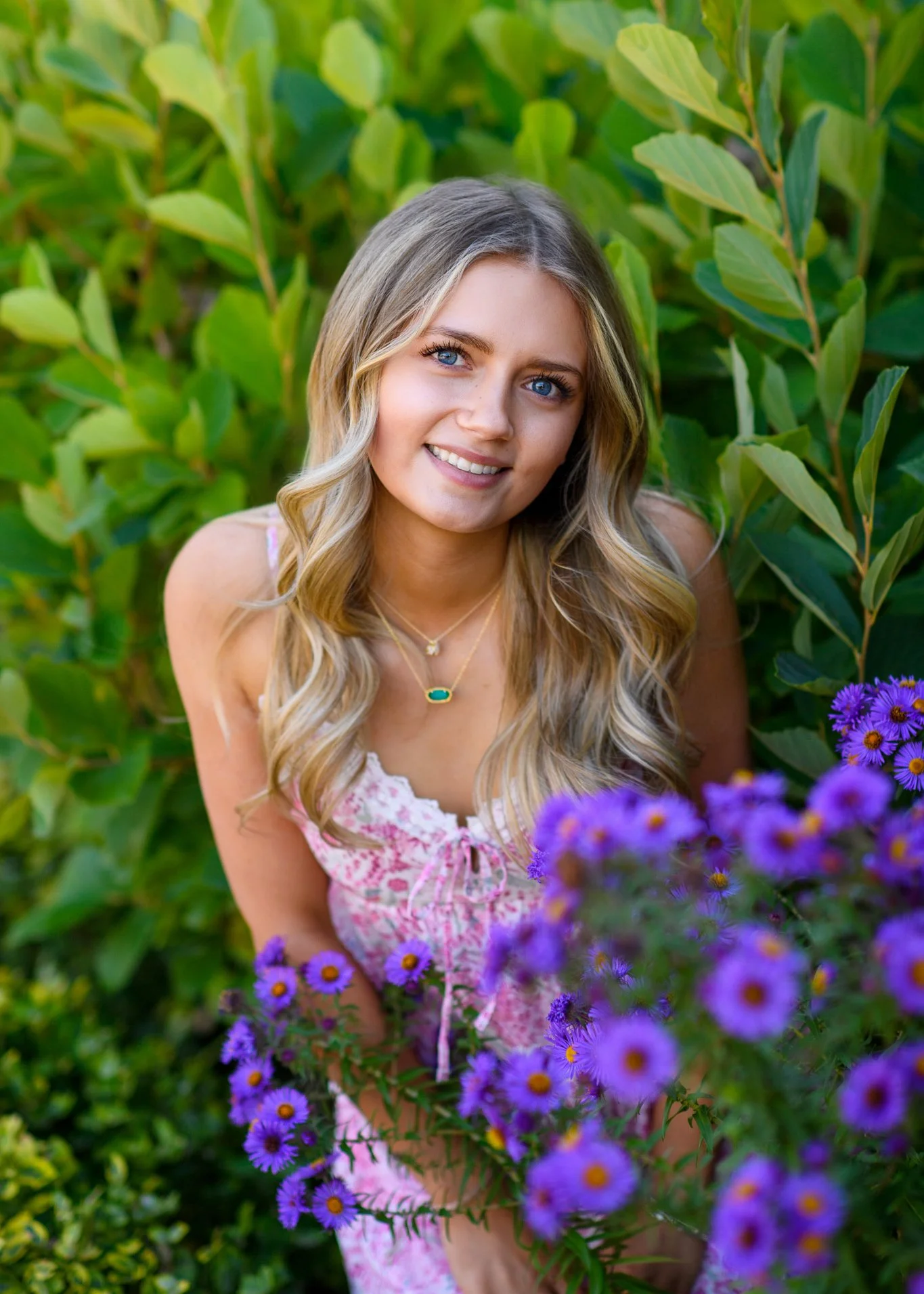 A young woman with long wavy blonde hair and blue eyes, smiling, outdoors among green foliage and purple flowers, wearing a pink and white dress and layered necklaces.