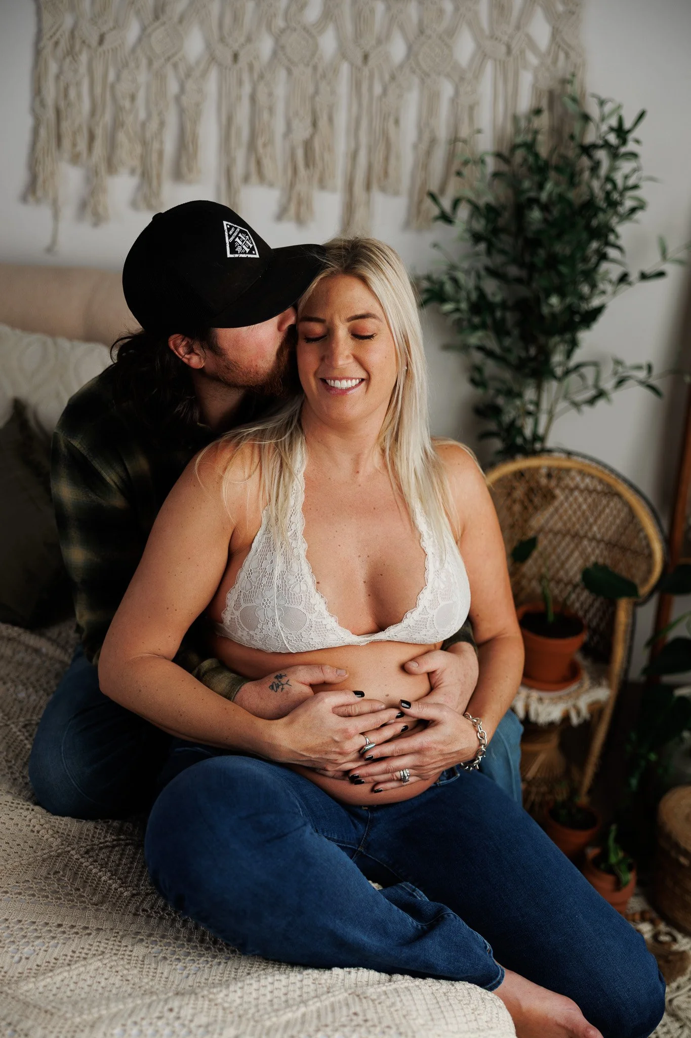 A man kissing a pregnant woman on her cheek while she smiles with her eyes closed, sitting on a bed in a cozy room decorated with plants and woven wall art.