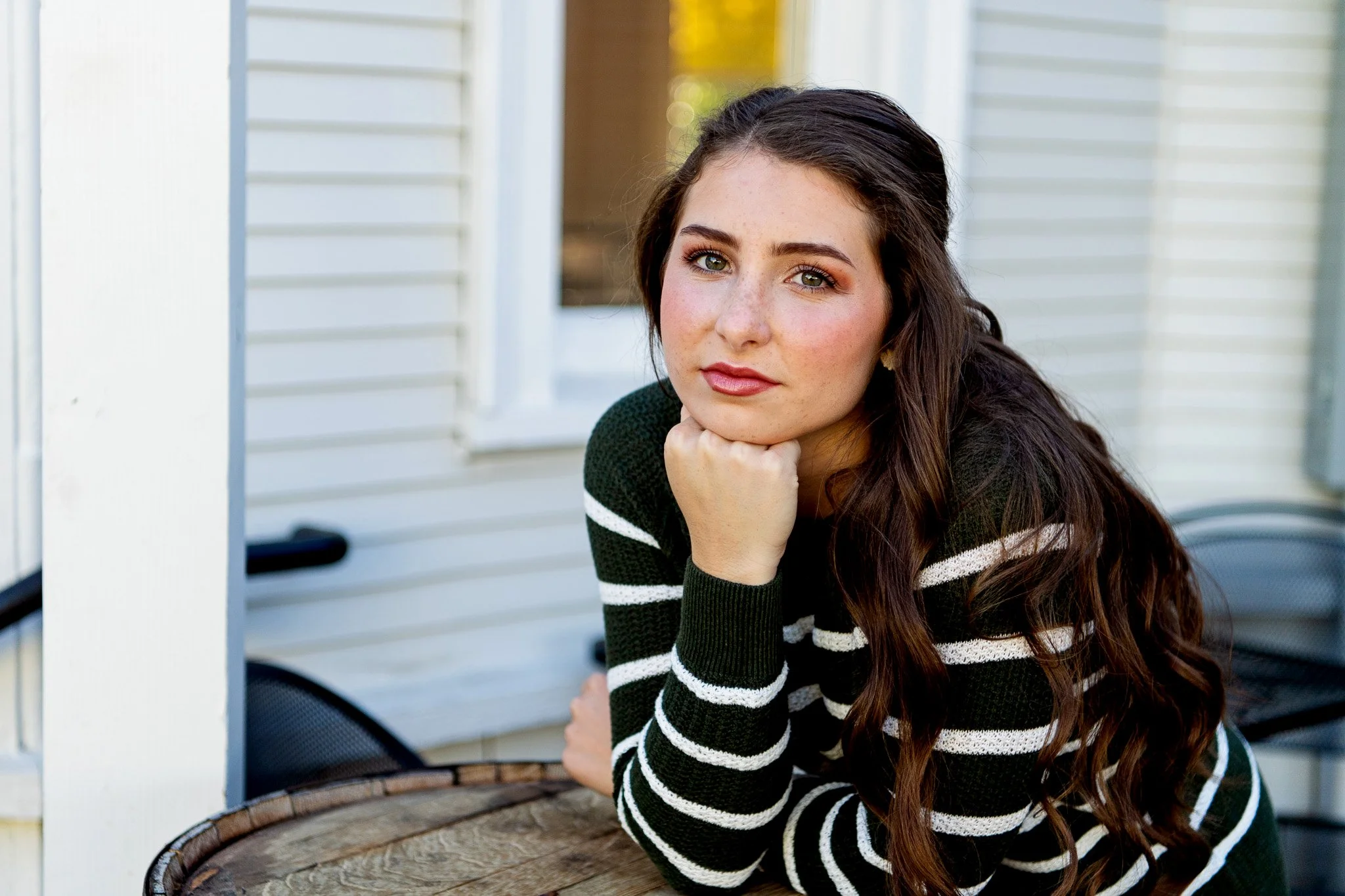 Young woman with long, wavy brown hair and freckles, resting her chin on her hand, wearing a dark green and white striped sweater, outdoors near a white building with a window.
