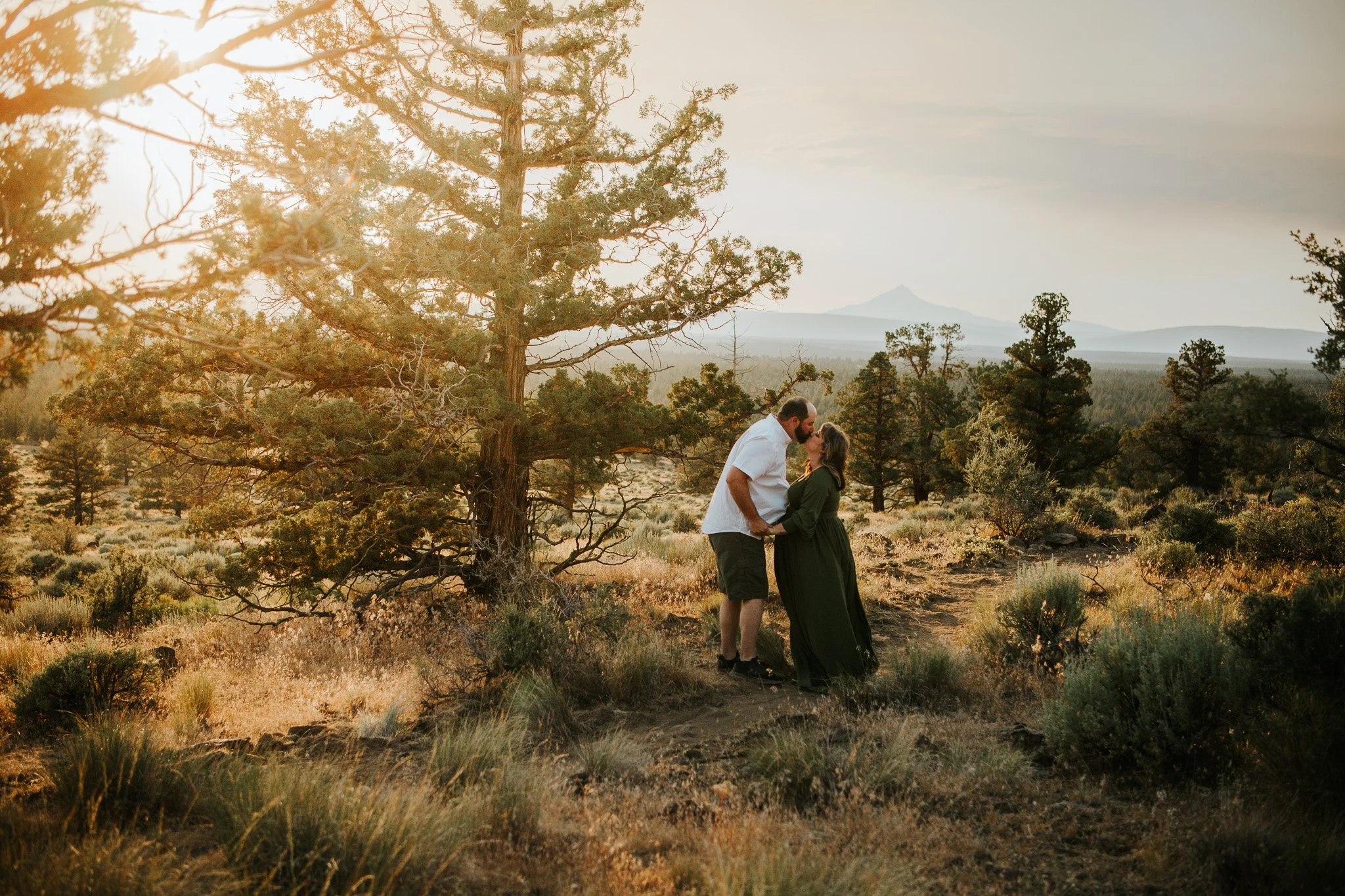 A couple stands close together on a trail in a desert landscape at sunset, surrounded by trees and bushes, with mountains in the background.