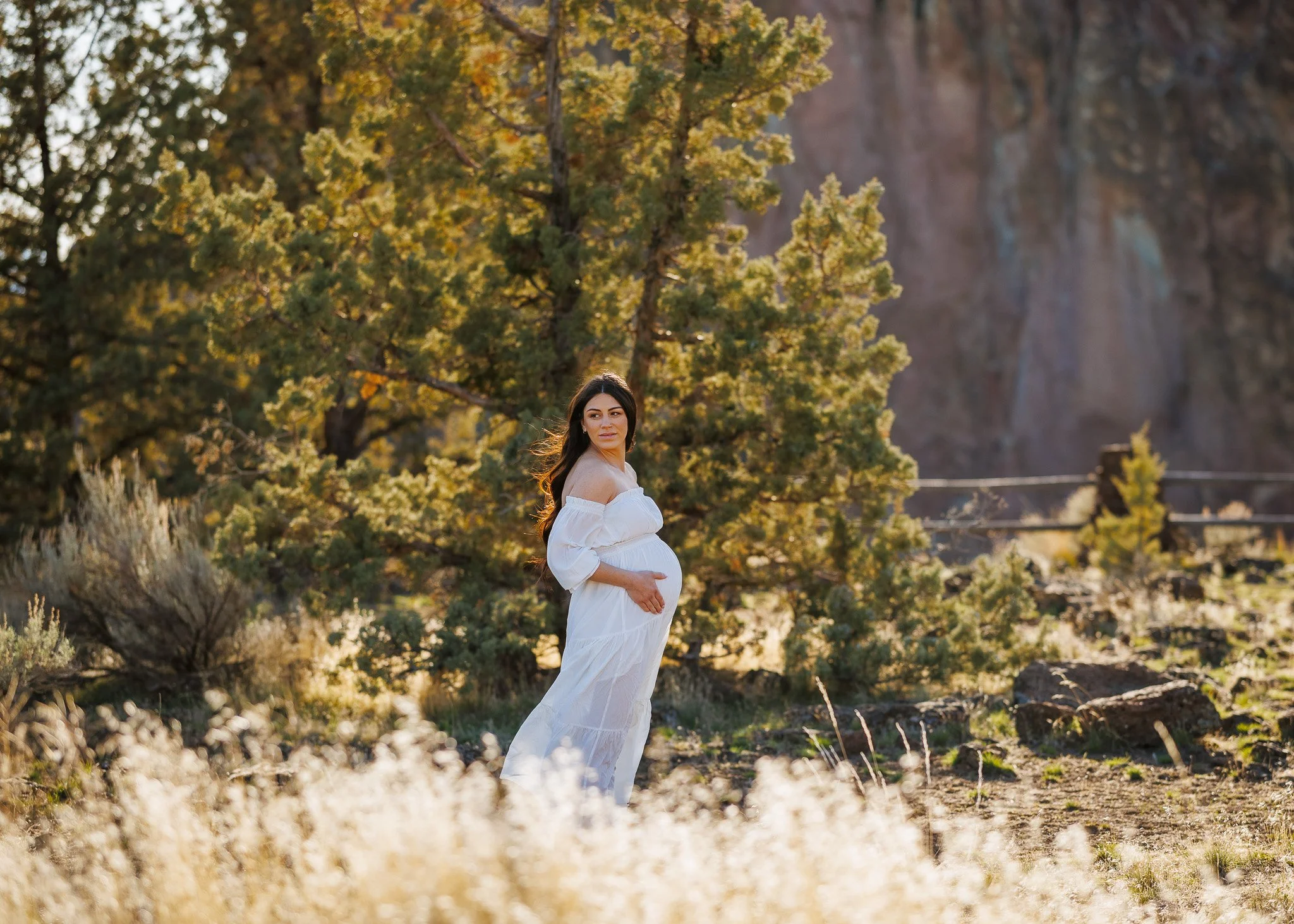Pregnant woman in a white dress standing outdoors among trees and rocks, with a mountain in the background, during sunset.