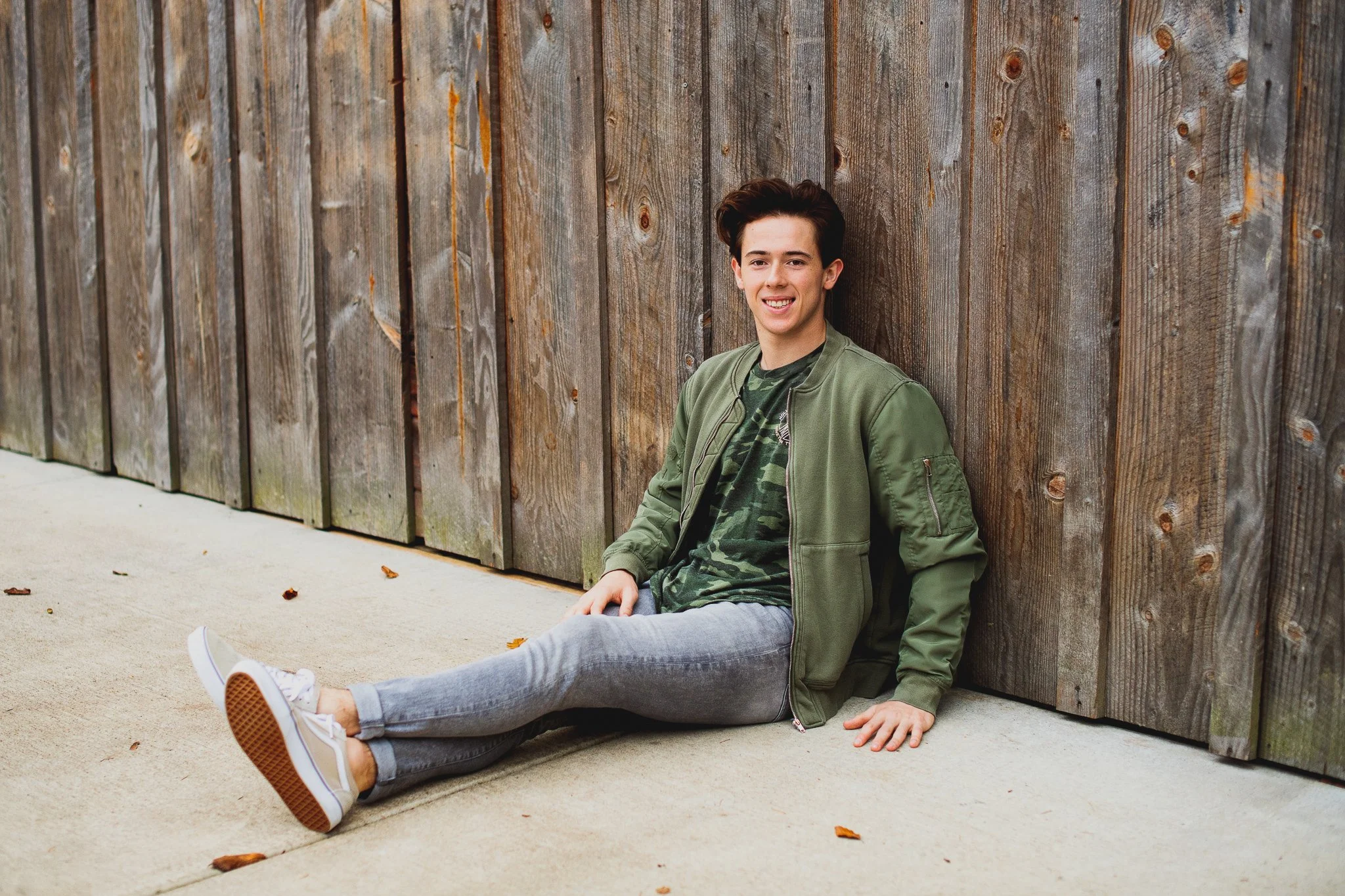 A young man sitting on the ground in front of a wooden fence, wearing a green bomber jacket, camouflage t-shirt, gray jeans, and white sneakers, smiling at the camera.