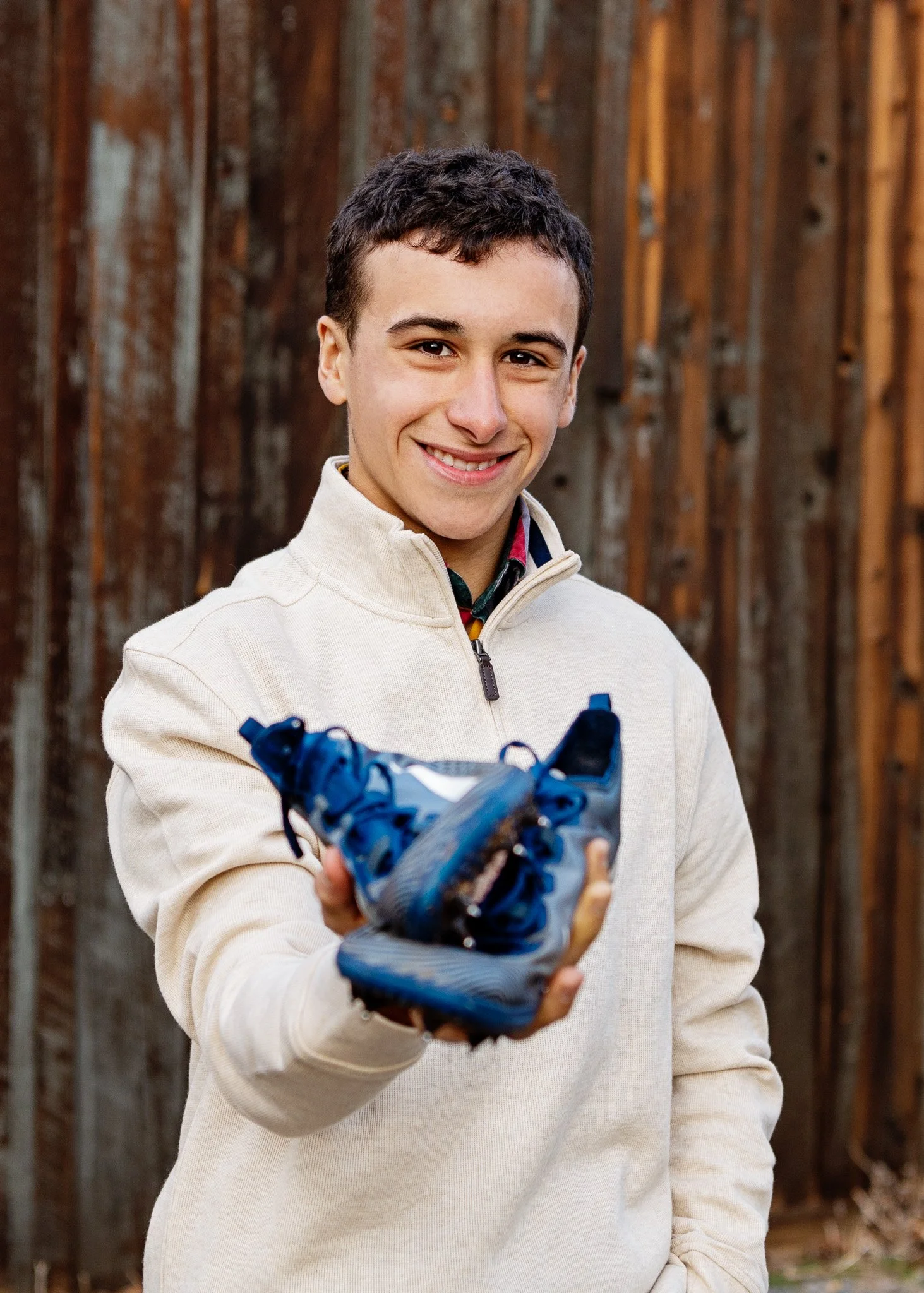 A young man with short dark hair is smiling and holding out a blue athletic shoe toward the camera outdoors, with a wooden fence in the background.