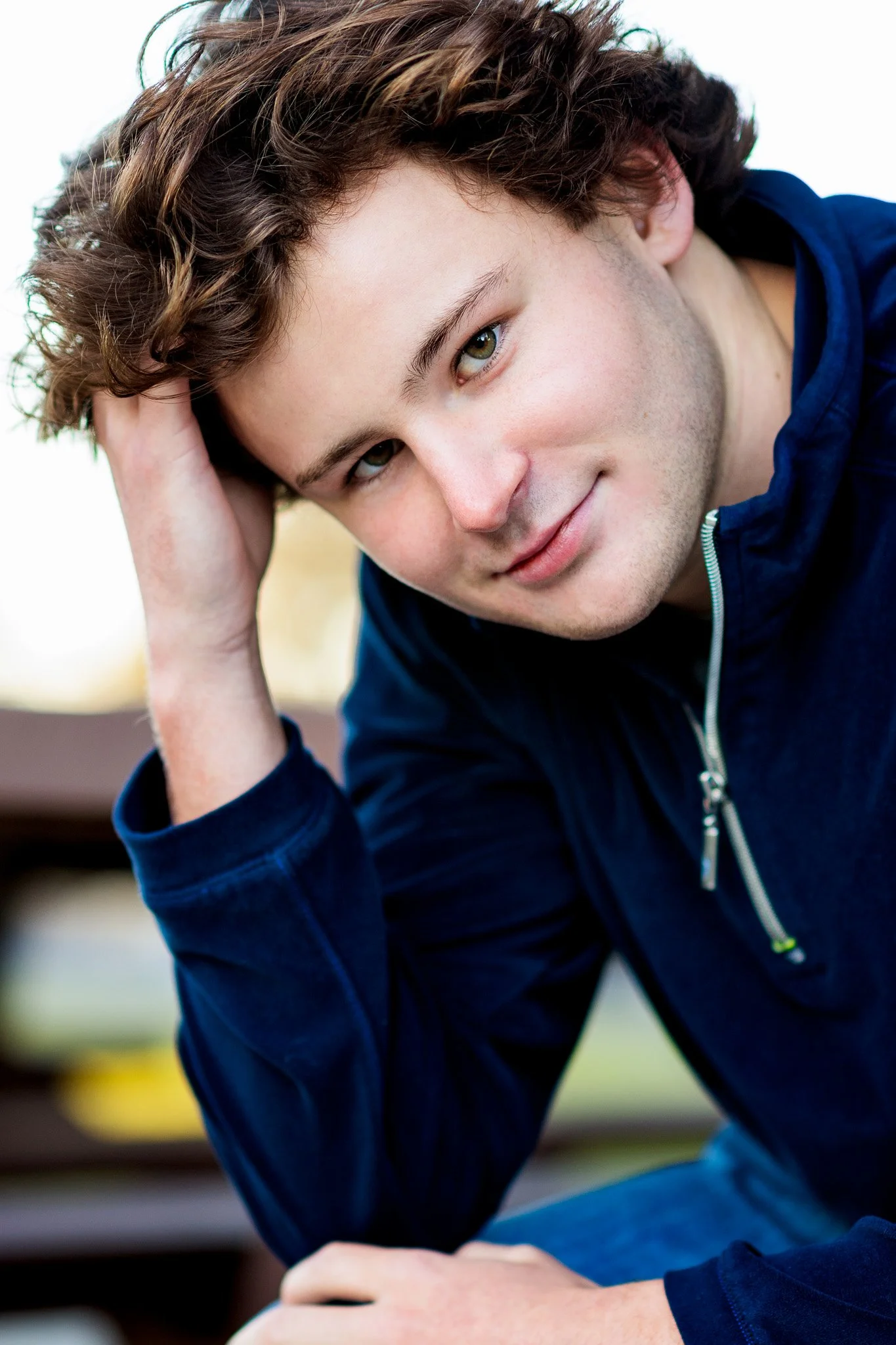Close-up of a young man with curly brown hair, green eyes, and light skin, resting his head on his hand, wearing a dark blue zip-up jacket, outdoors during daytime.