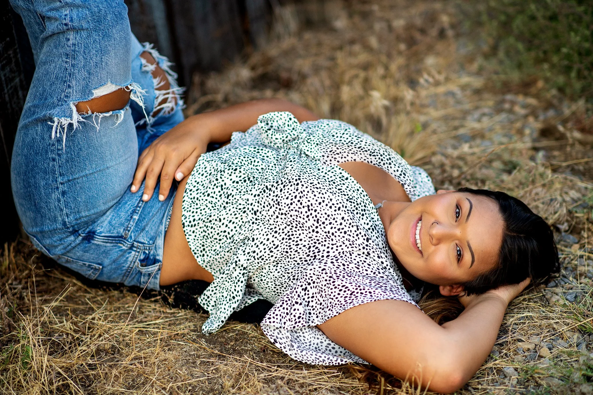A woman lying on the ground outdoors, smiling, with her head resting on her arm, wearing a white polka dot top and ripped jeans.