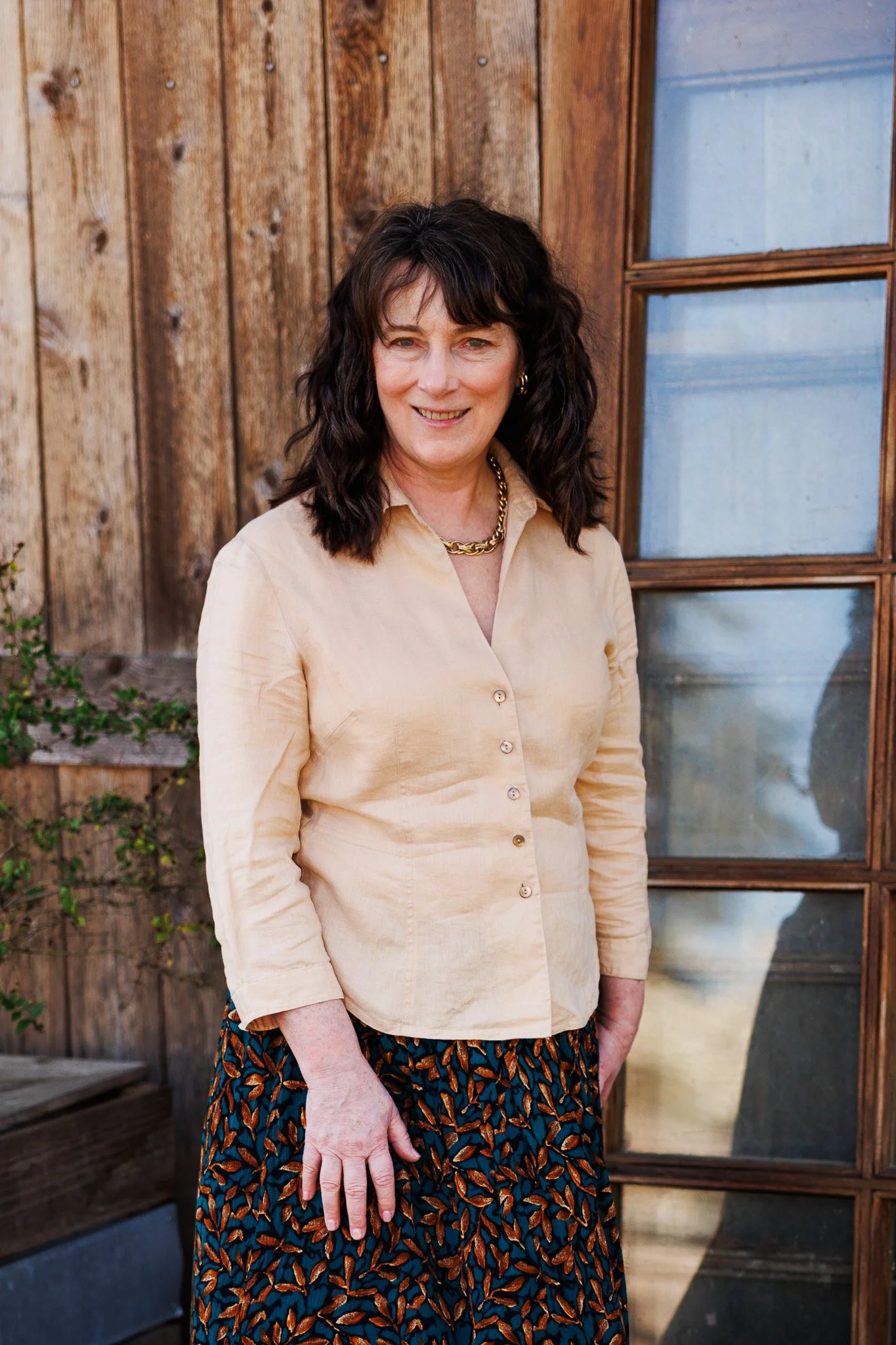 A woman with dark, curly hair standing outdoors in front of a wooden wall with a window. She is wearing a light beige button-up shirt, patterned skirt, and gold jewelry.