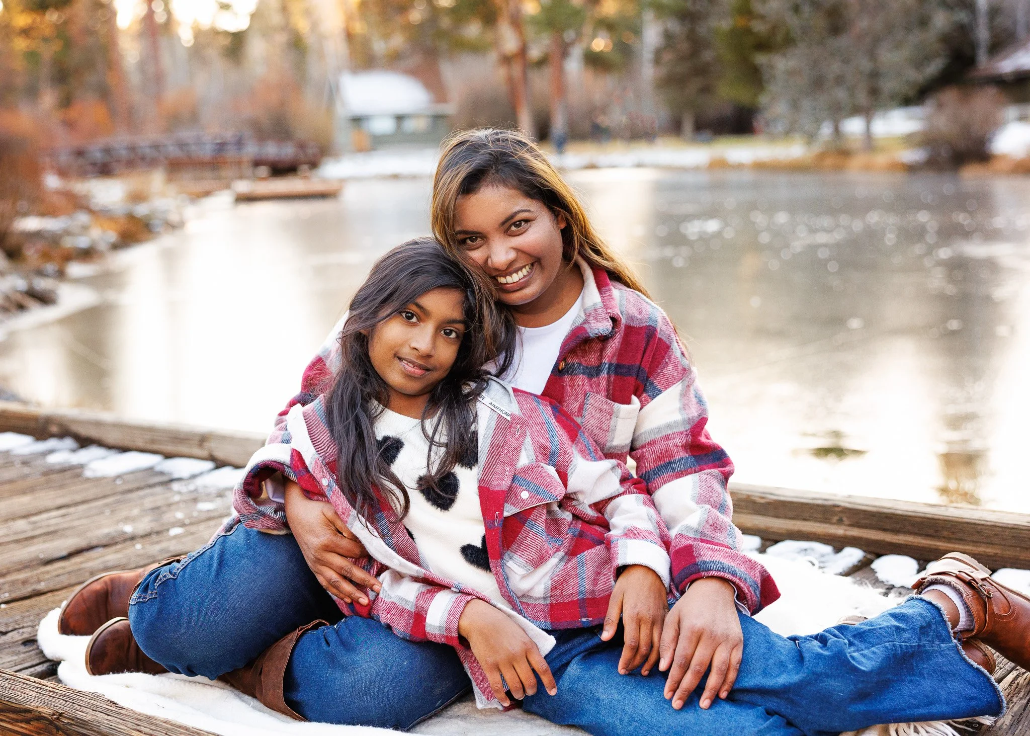 A woman and her daughter sitting on a wooden dock by a river, smiling, with fall trees and snow in the background.
