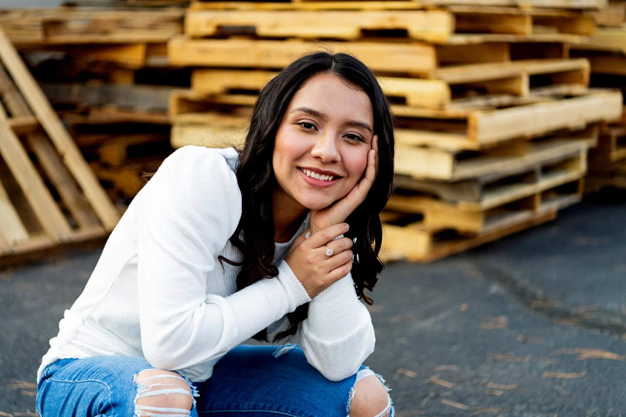 A young woman with long dark hair and a white sweater sitting outdoors, resting her face on her hand, smiling at the camera with a background of stacked wooden pallets.