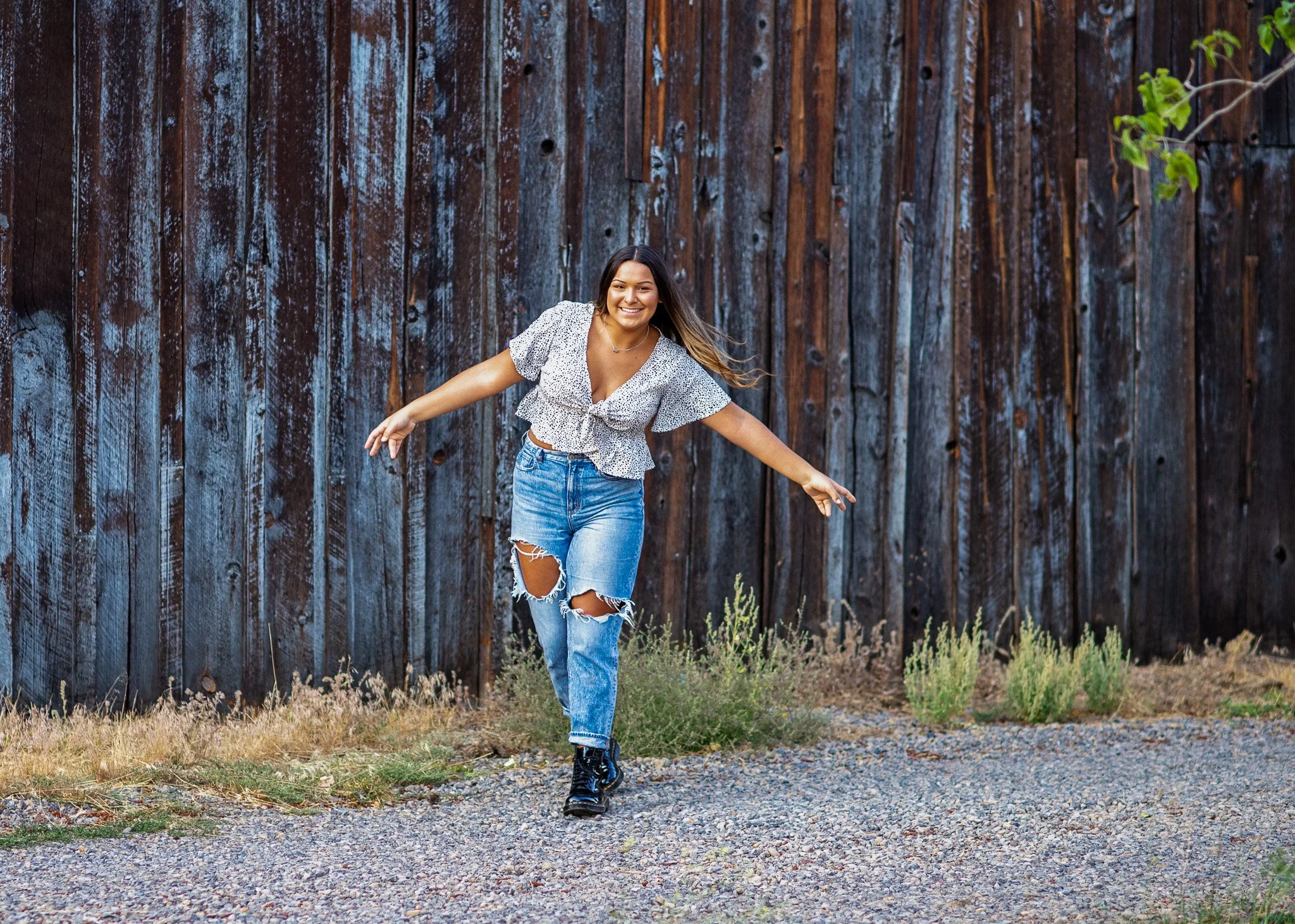 A woman with long hair wearing a white patterned crop top, ripped jeans, and black shoes, smiling and balancing on one foot, outdoors in front of a tall, weathered wooden fence with some greenery.