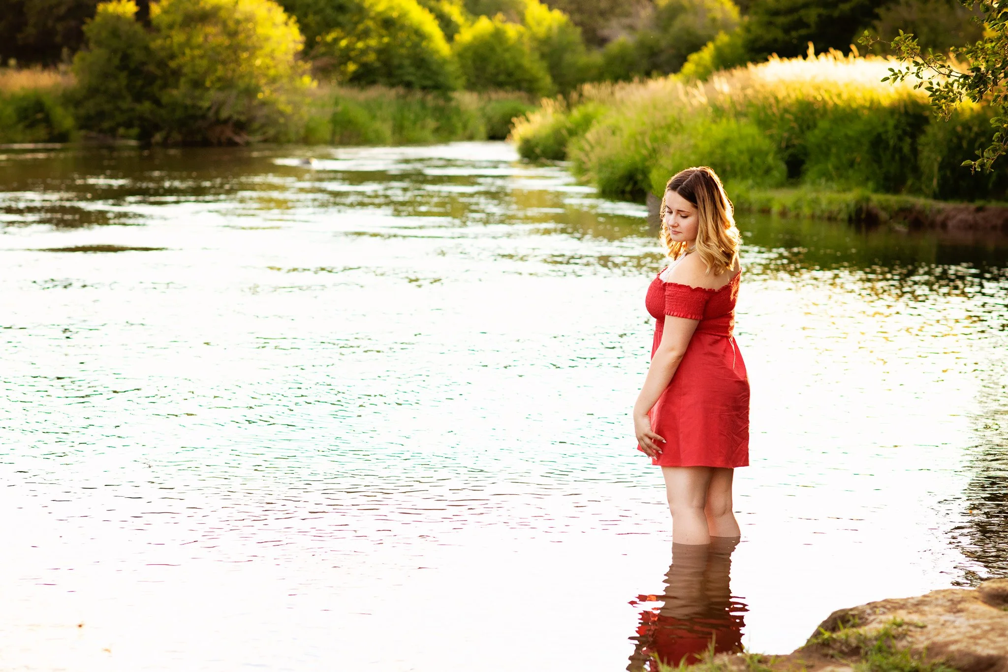 A young woman in a red dress standing in a river during sunset with greenery and trees in the background.