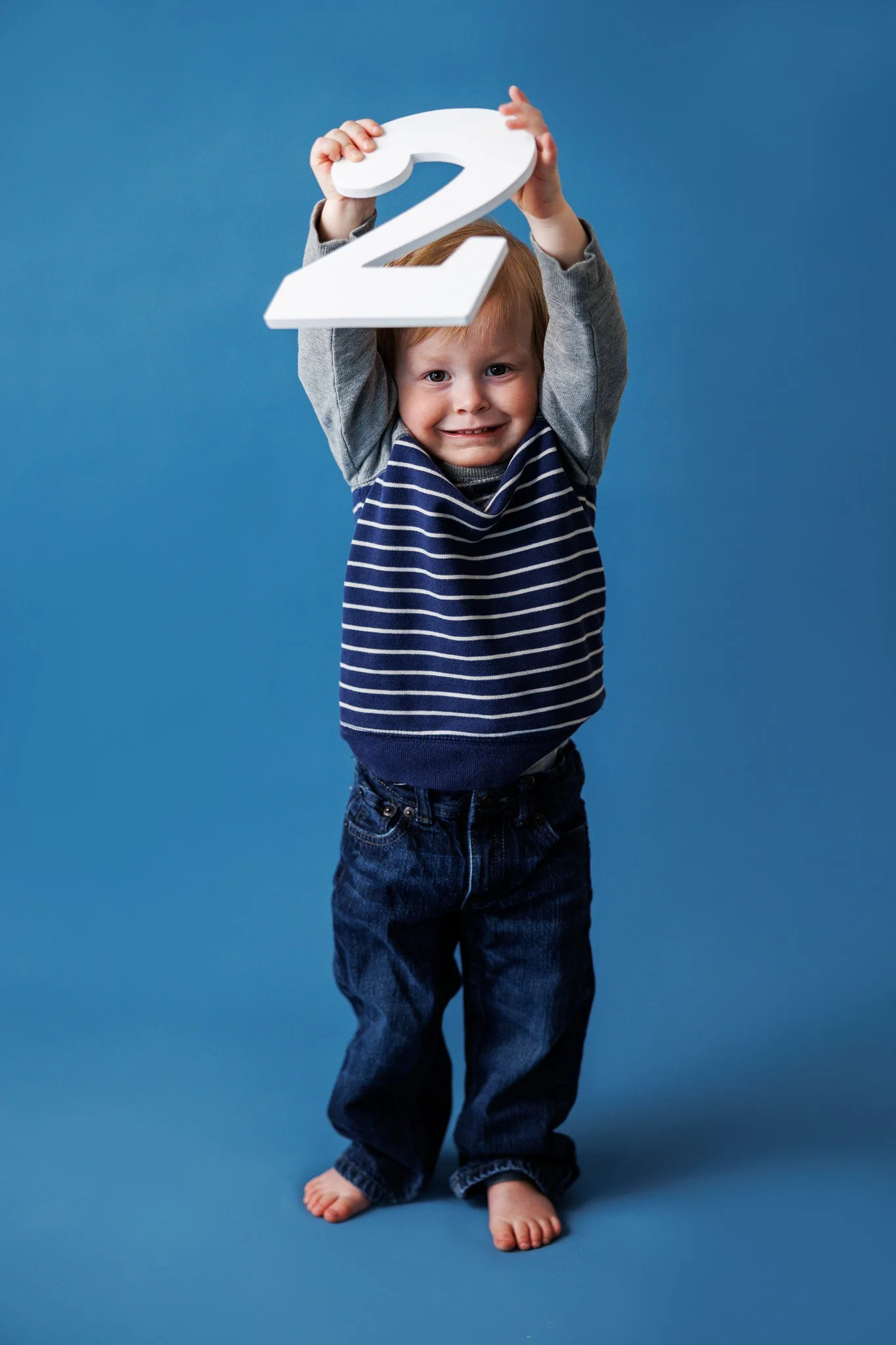 A young child standing barefoot on a blue background, smiling and holding a large white number 2 above his head.