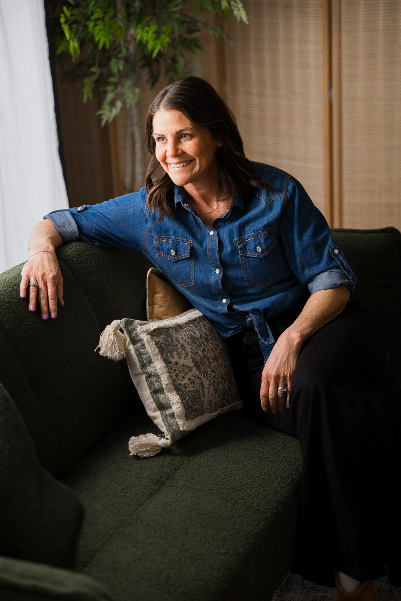 A woman with shoulder-length dark hair smiling and sitting on a dark green sofa, leaning on her left arm, with a decorative pillow beside her, in a cozy living room setting.