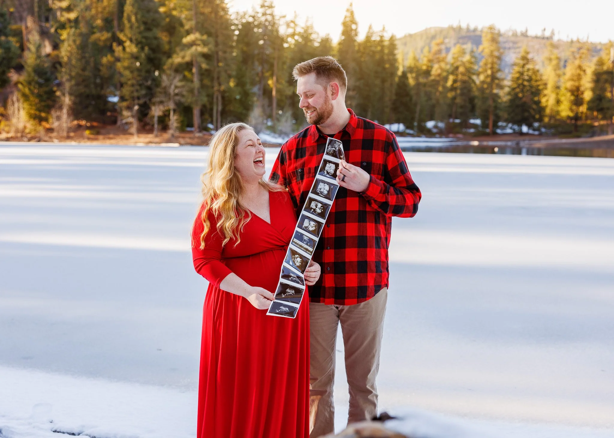 A pregnant woman in a red dress and a man in a red and black plaid shirt smile at each other outdoors on a snowy lake with a forest and mountains in the background, holding ultrasound pictures.