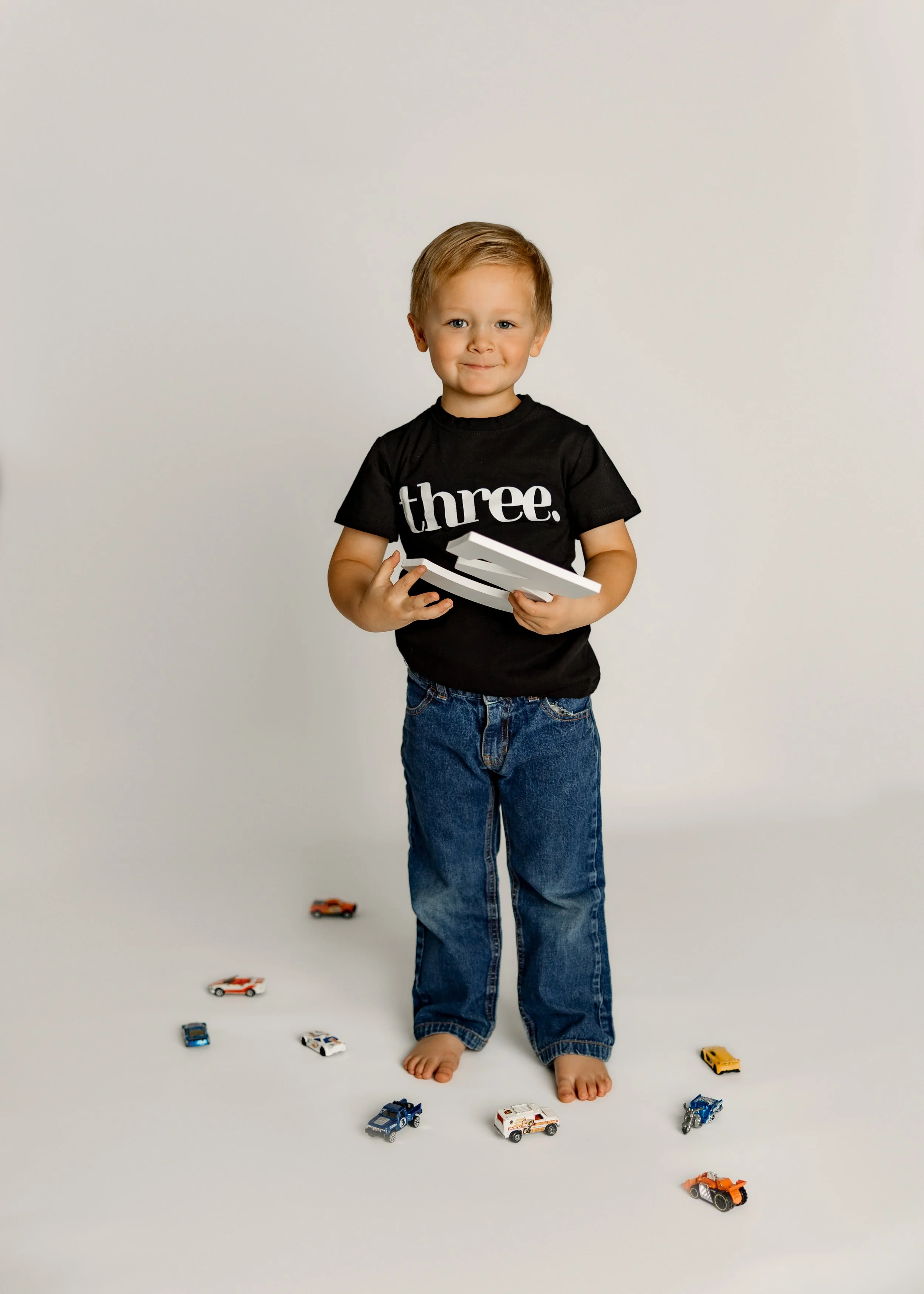 A young boy standing barefoot on a white background, holding a few books, wearing a black t-shirt with the word 'three.' printed on it, and blue jeans, surrounded by toy cars on the floor.