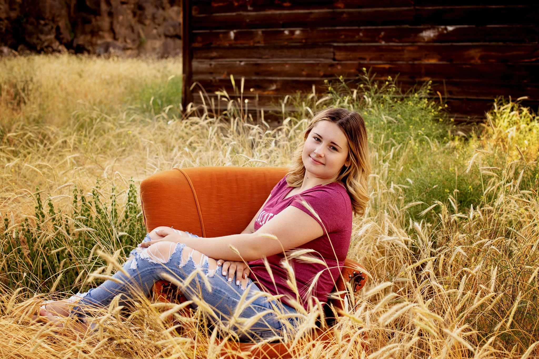A young woman with blonde hair and a pink T-shirt sitting on an orange armchair in a field of tall dried grass, with a wooden structure in the background.