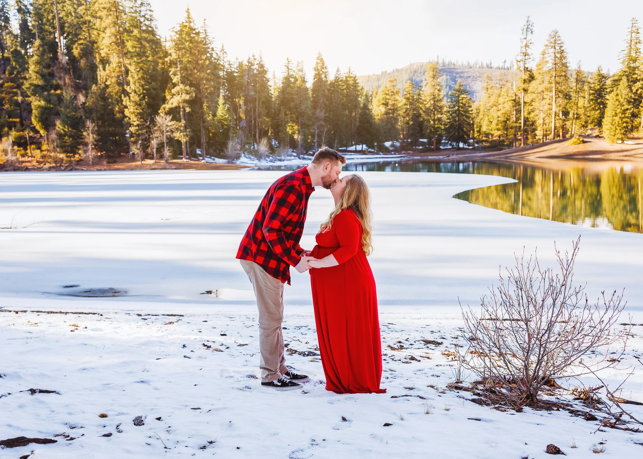A couple holding hands and kissing in front of a snow-covered lakeside with pine trees and mountains in the background.