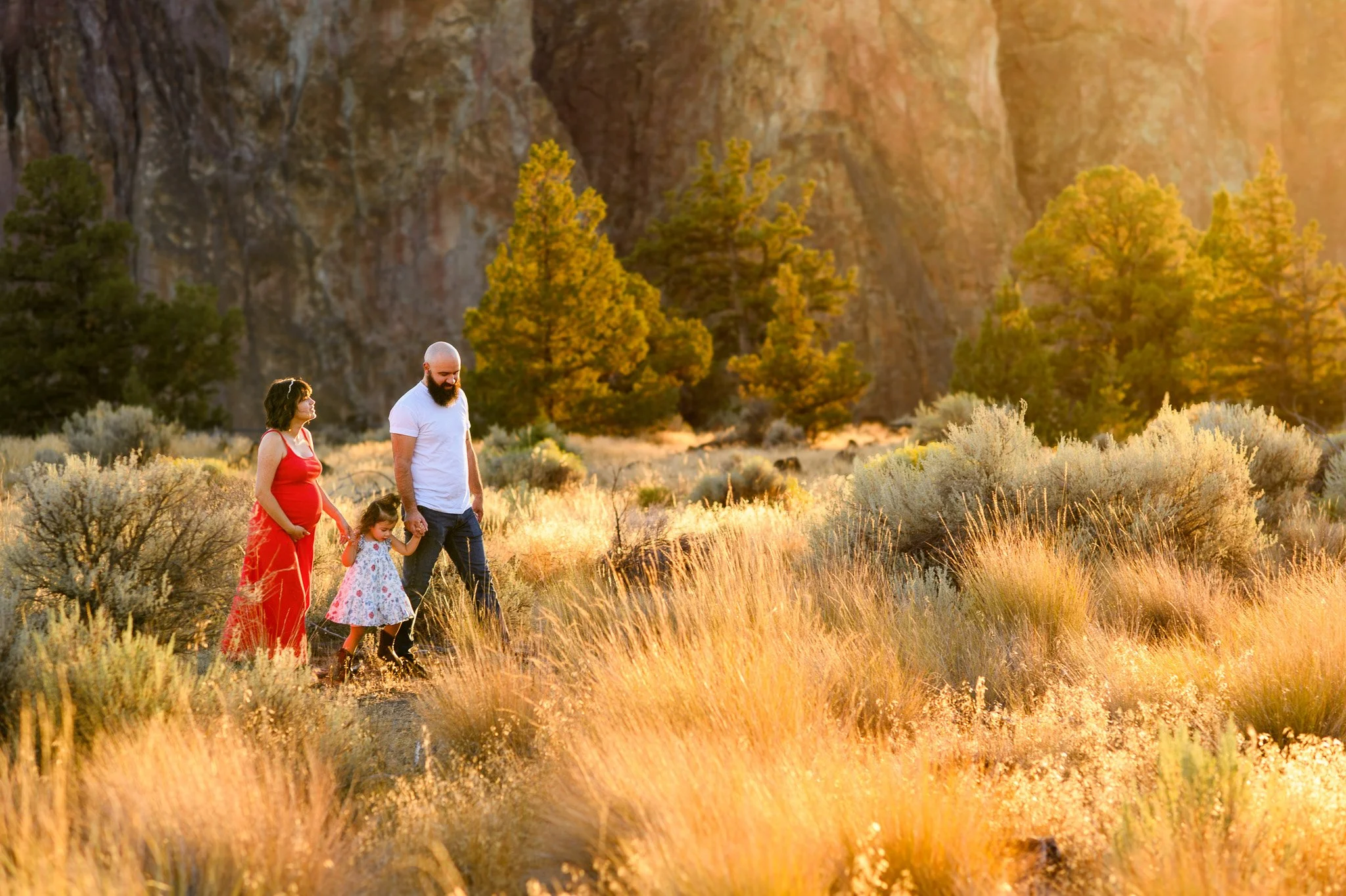 A family of three walking through a field of tall grass and shrubs during sunset, with trees and rocky cliffs in the background.